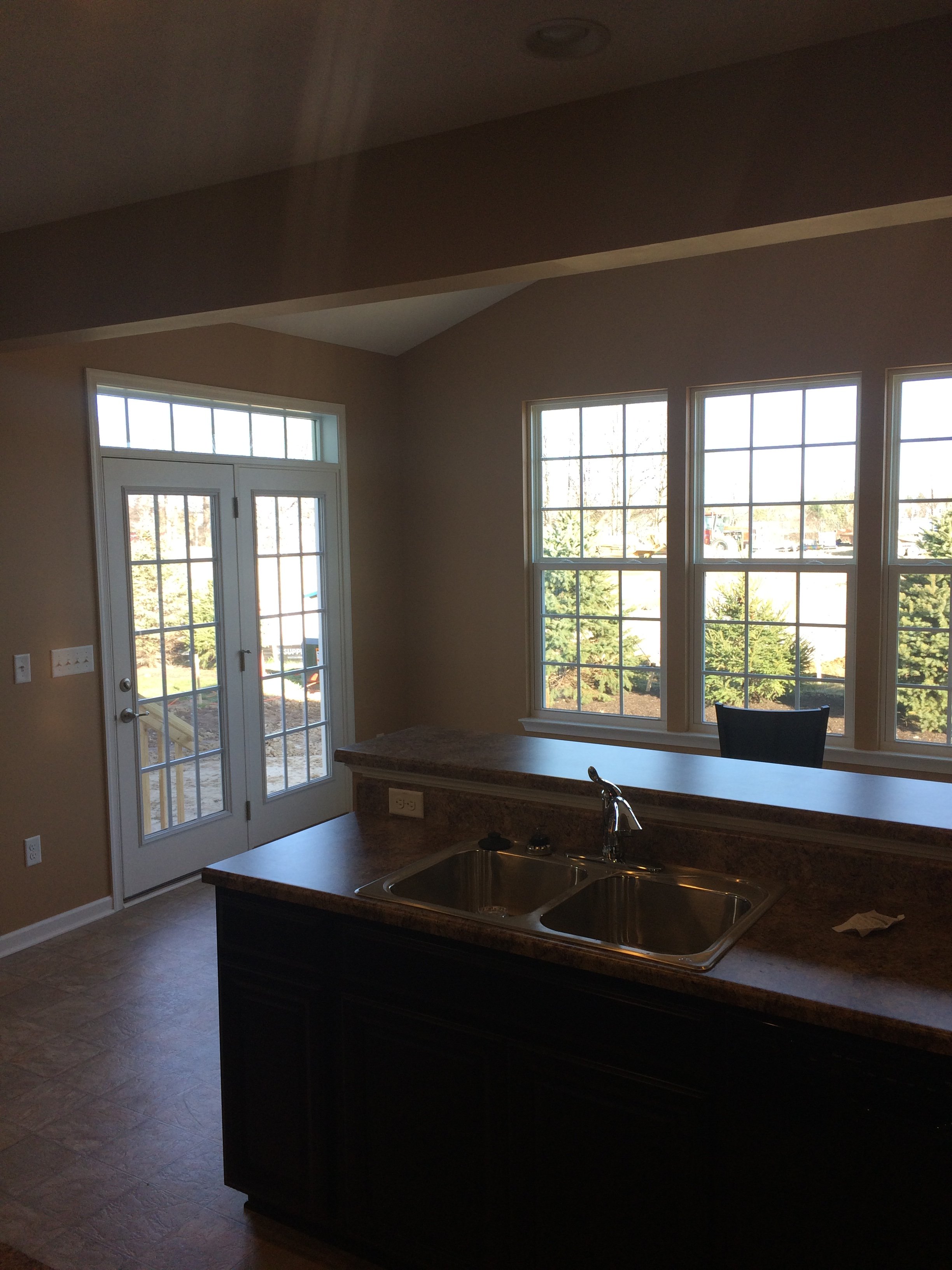 Interior of a kitchen with large windows, a dark kitchen island with a double sink, and a view of trees outside.