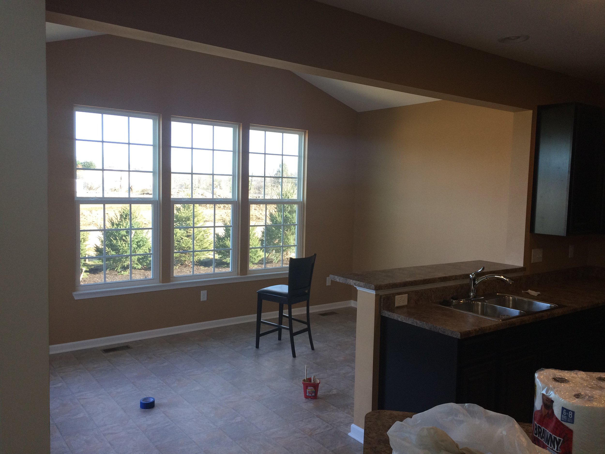 The image shows a kitchen with large windows, beige walls, and a granite countertop. There are a few chairs, a bucket, and some construction supplies indicating ongoing renovation or cleaning.