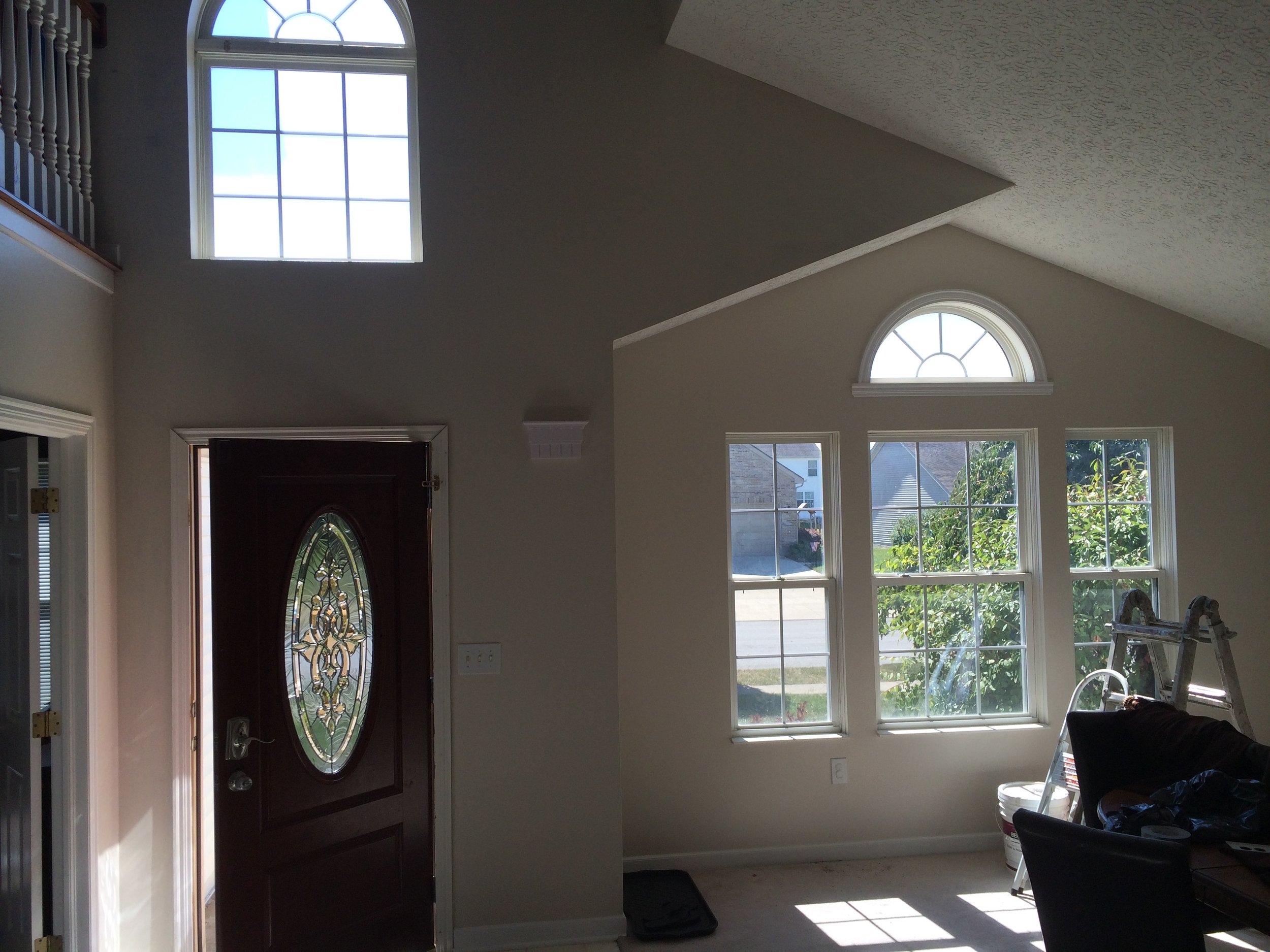 Interior view of a living room with large windows, one with an arched top, allowing in natural light, and a front door with a decorative glass panel. Some furniture and items are visible near the bottom right corner.