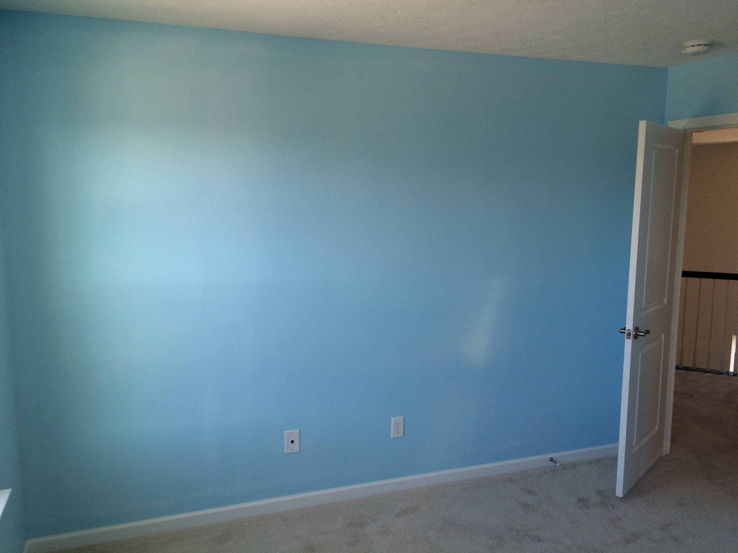 A room with light blue painted walls, a partially open door, and multiple electrical outlets along the wall. The floor is carpeted, and there is a smoke detector on the ceiling.