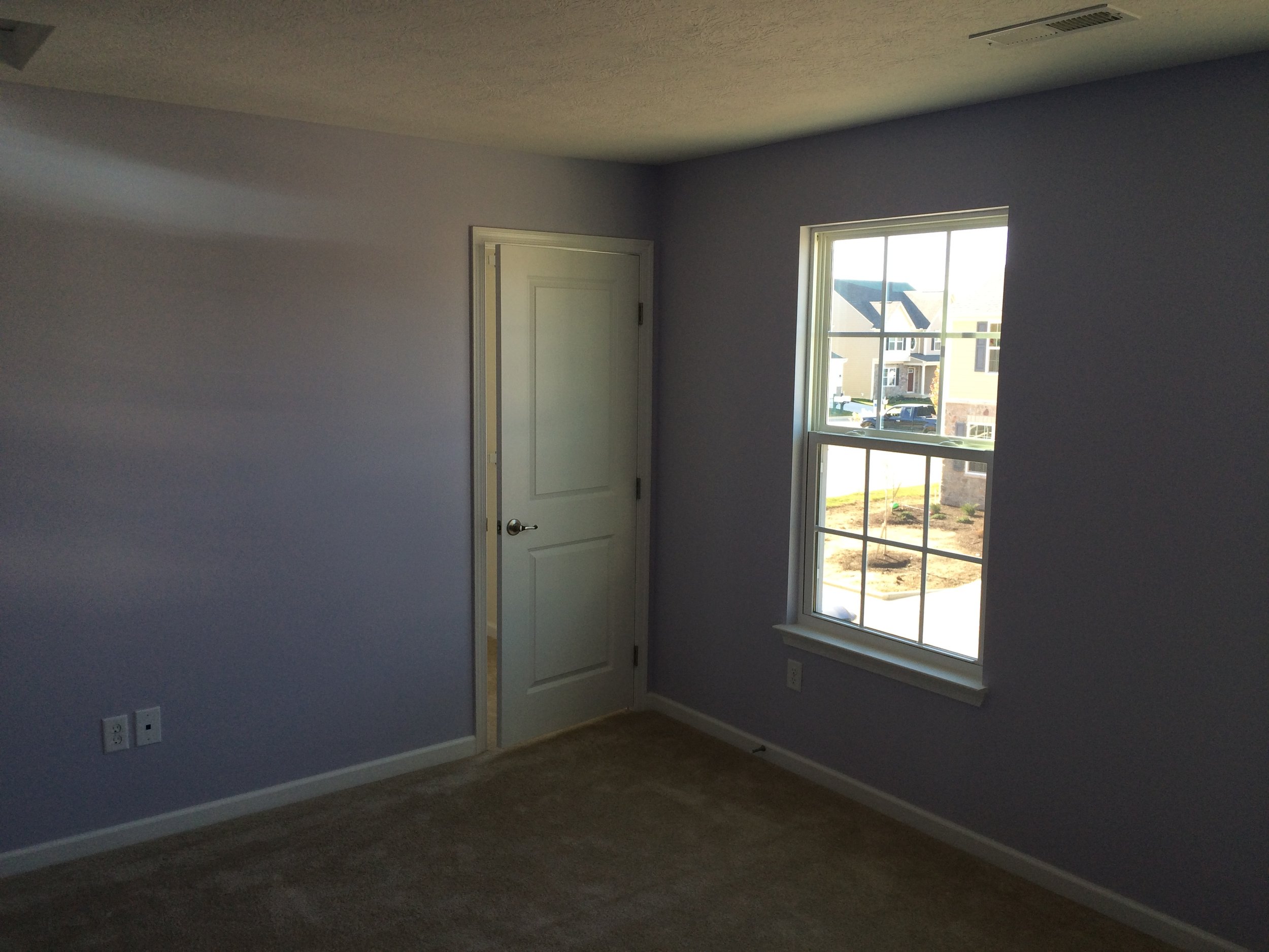 Empty room with a large window showing a suburban neighborhood, a closed white door, and carpeted flooring.