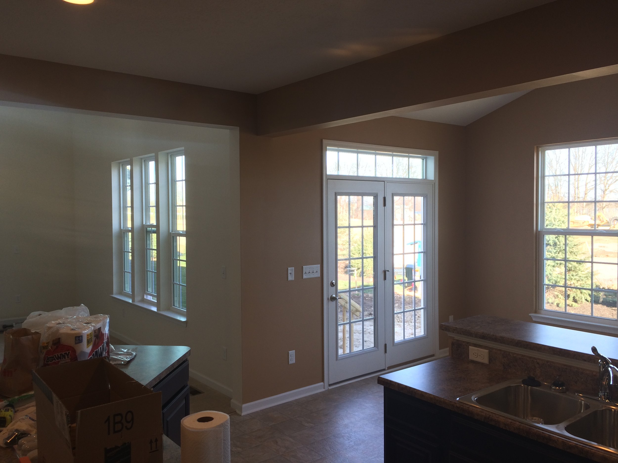 Interior view of a room with large windows and a glass door, sunlight streaming in, beige walls, and a kitchen counter on the right. Packing boxes and supplies are on the left.