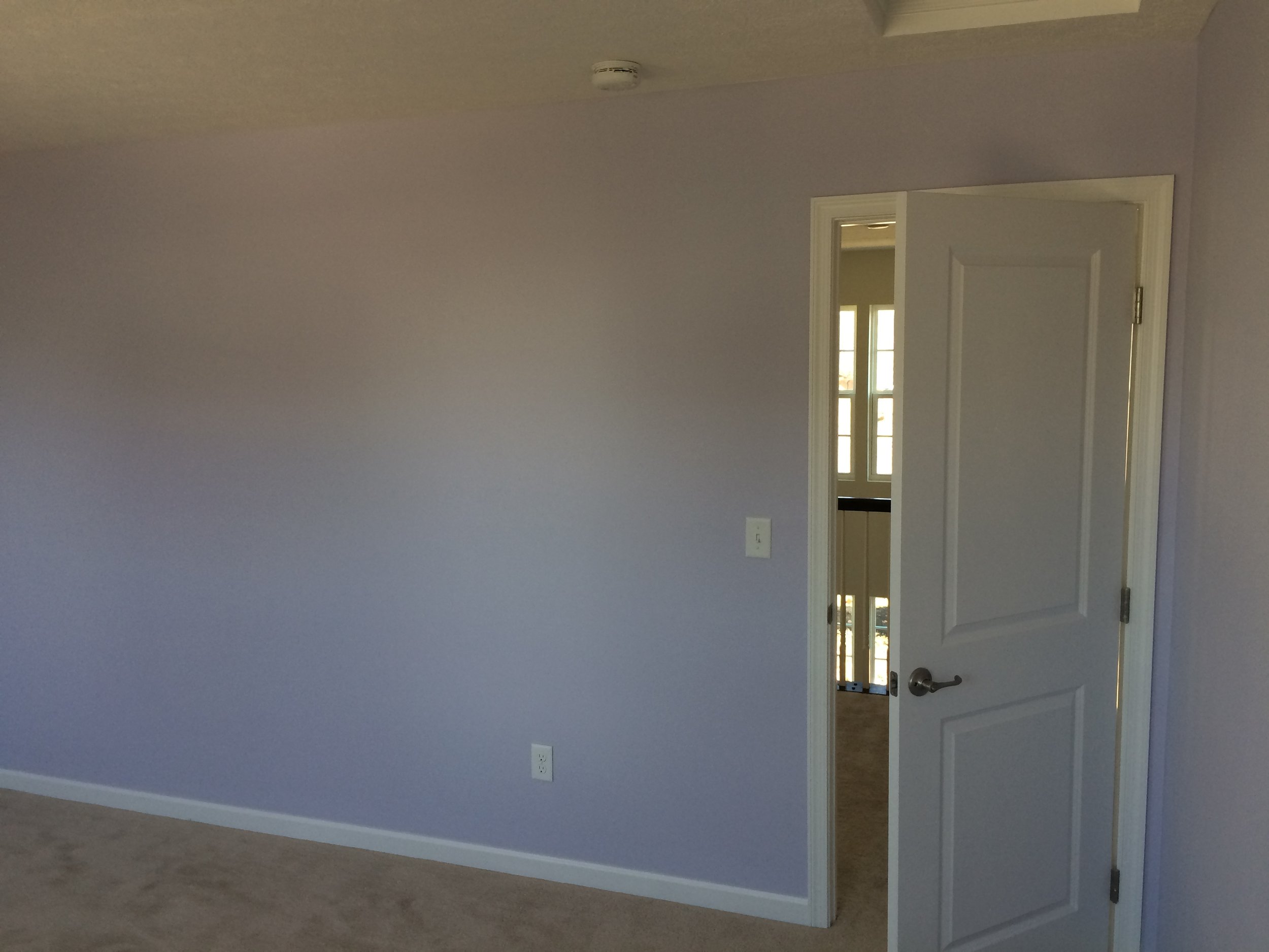 An empty room with gray walls, carpeted floor, an open door leading to a hallway, and a ceiling smoke detector