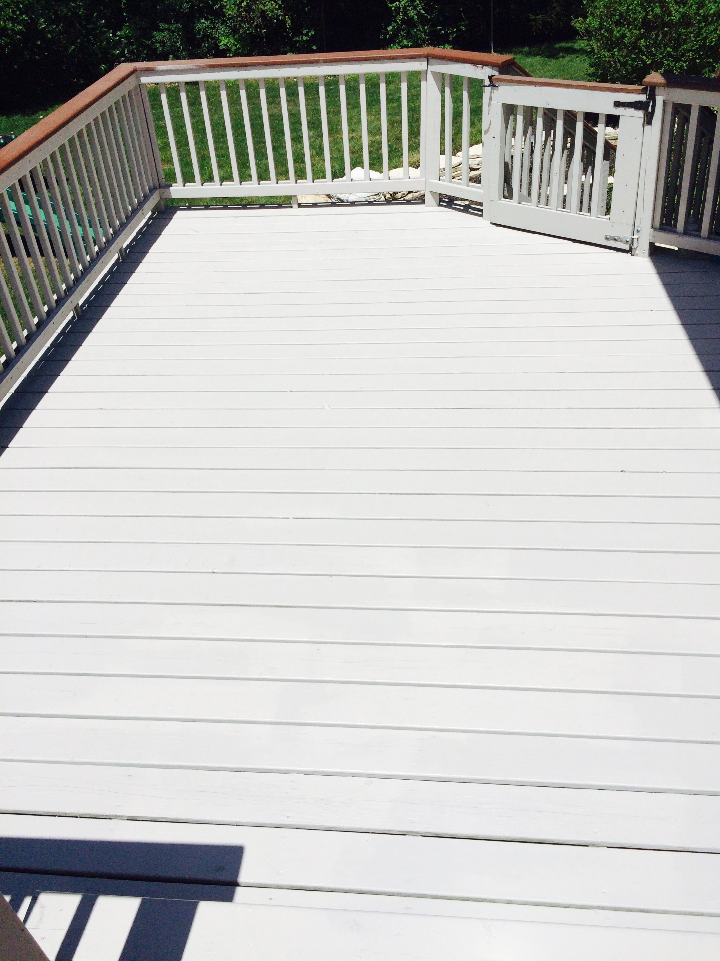 A white wooden deck with railings, some greenery and trees in the background, and a shadow of a chair or table in the foreground.
