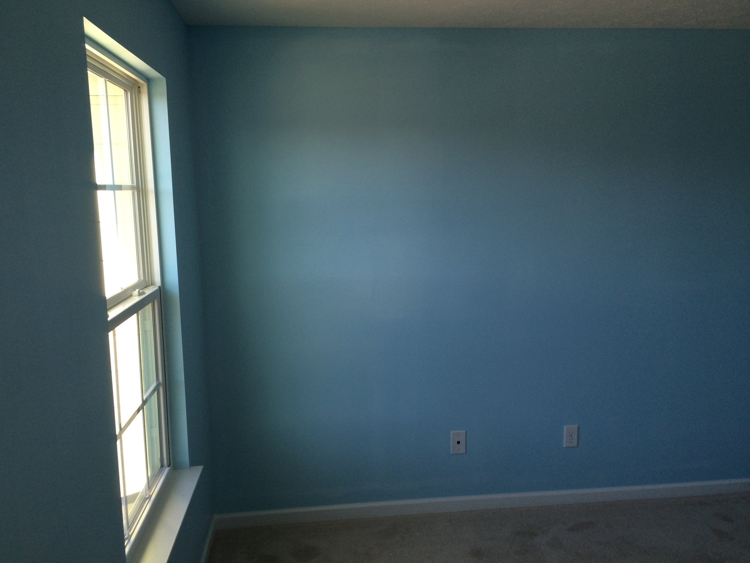 Empty blue room with a window on the left and three electrical outlets on the wall.