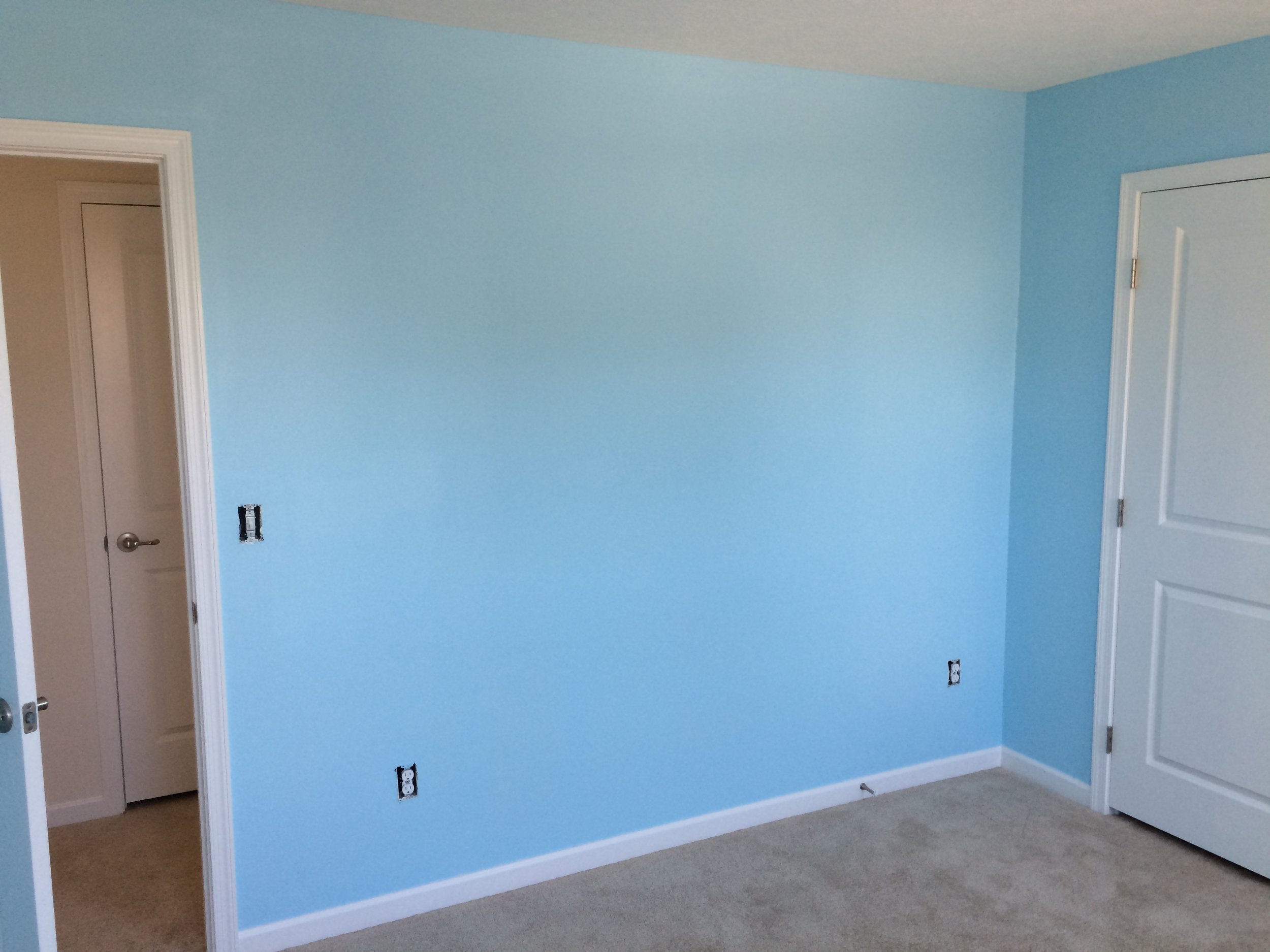 Empty room with blue walls, white baseboards, and door frames. There are multiple electrical outlets and a switch box on the wall, with two doors—one slightly open, revealing a beige room inside.