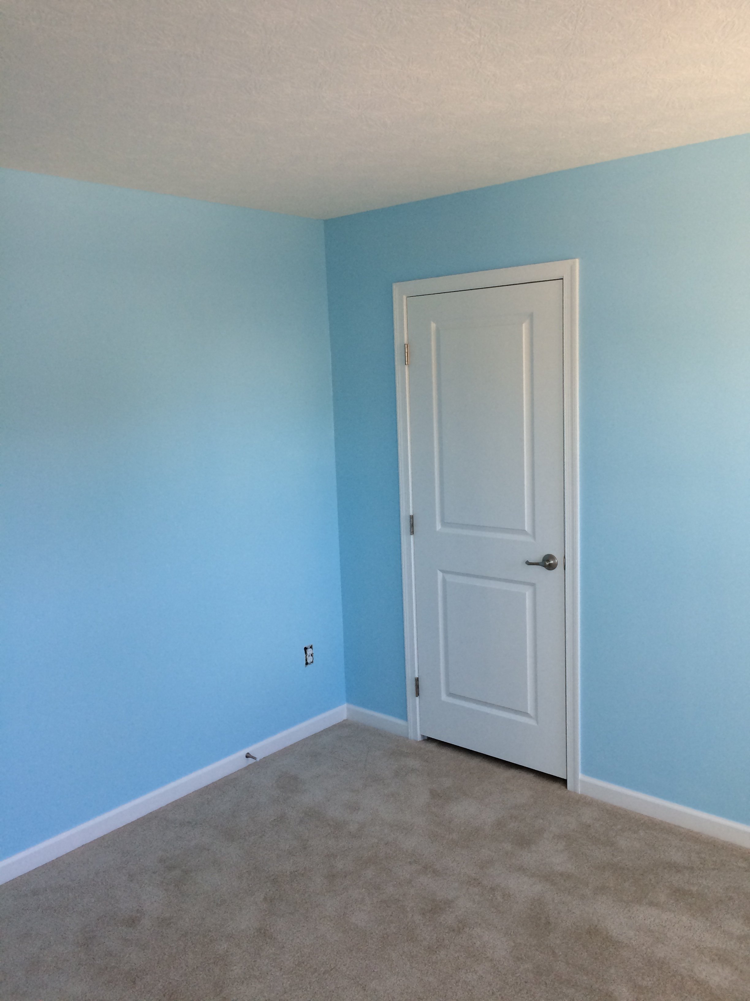 A room with light blue walls, a white door, beige carpet, and an electrical outlet on the wall.
