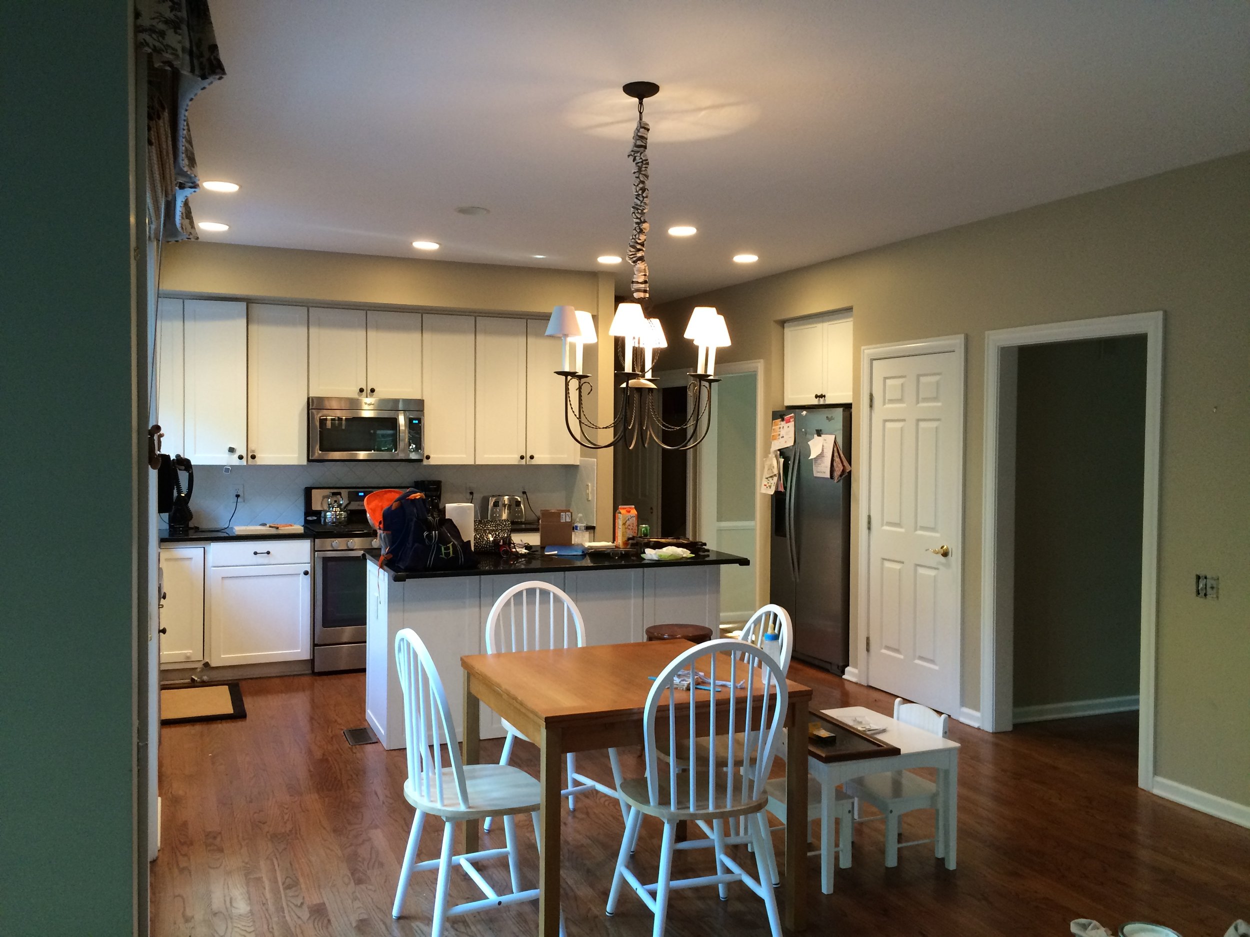 A kitchen and dining area with a wooden dining table with four white chairs, a white kitchen with stainless steel appliances, overhead lighting, a chandelier above the table, and various items on the counter and table.