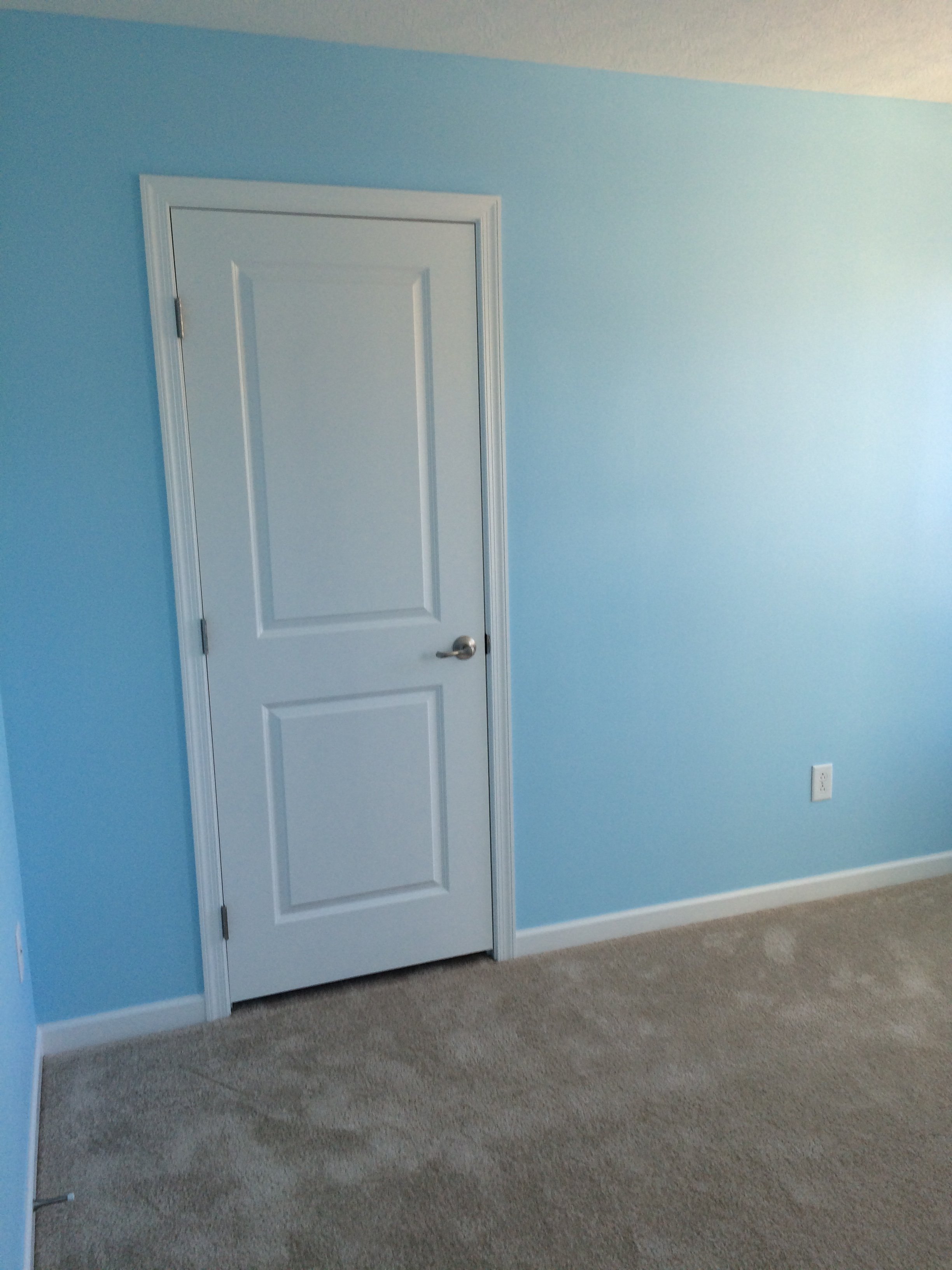 A room with blue walls, beige carpet, a white door with a round doorknob, and an electrical outlet near the floor.