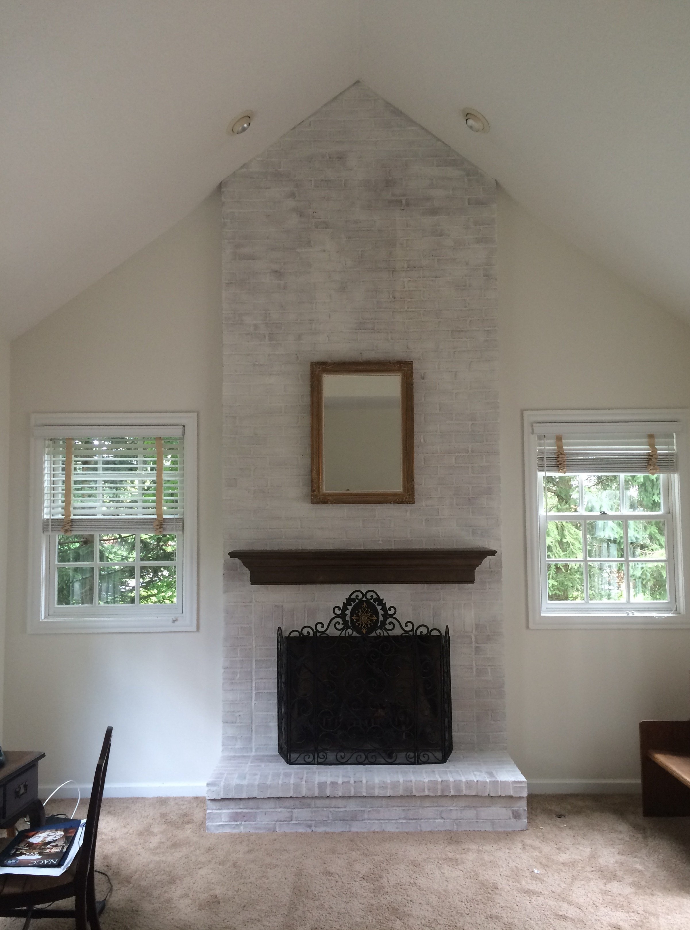 A living room with a white brick fireplace, two windows on either side, and a mirror above the fireplace. There is a brown carpet and some furniture including a black metal fireplace screen and a partial view of a chair and table.