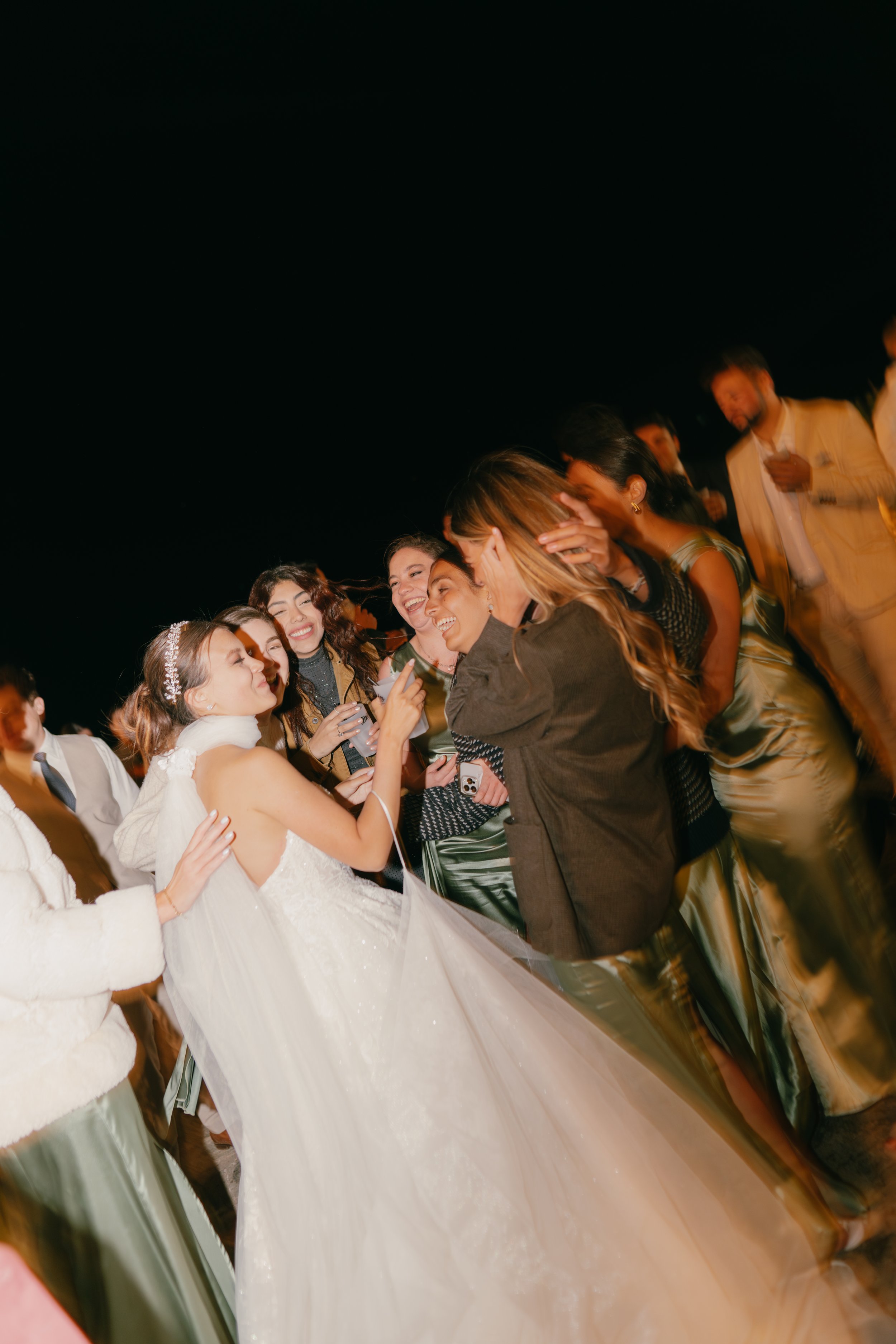 Group of women at a wedding celebration, with the bride in a white wedding dress and veil, laughing and taking photos with friends at night.