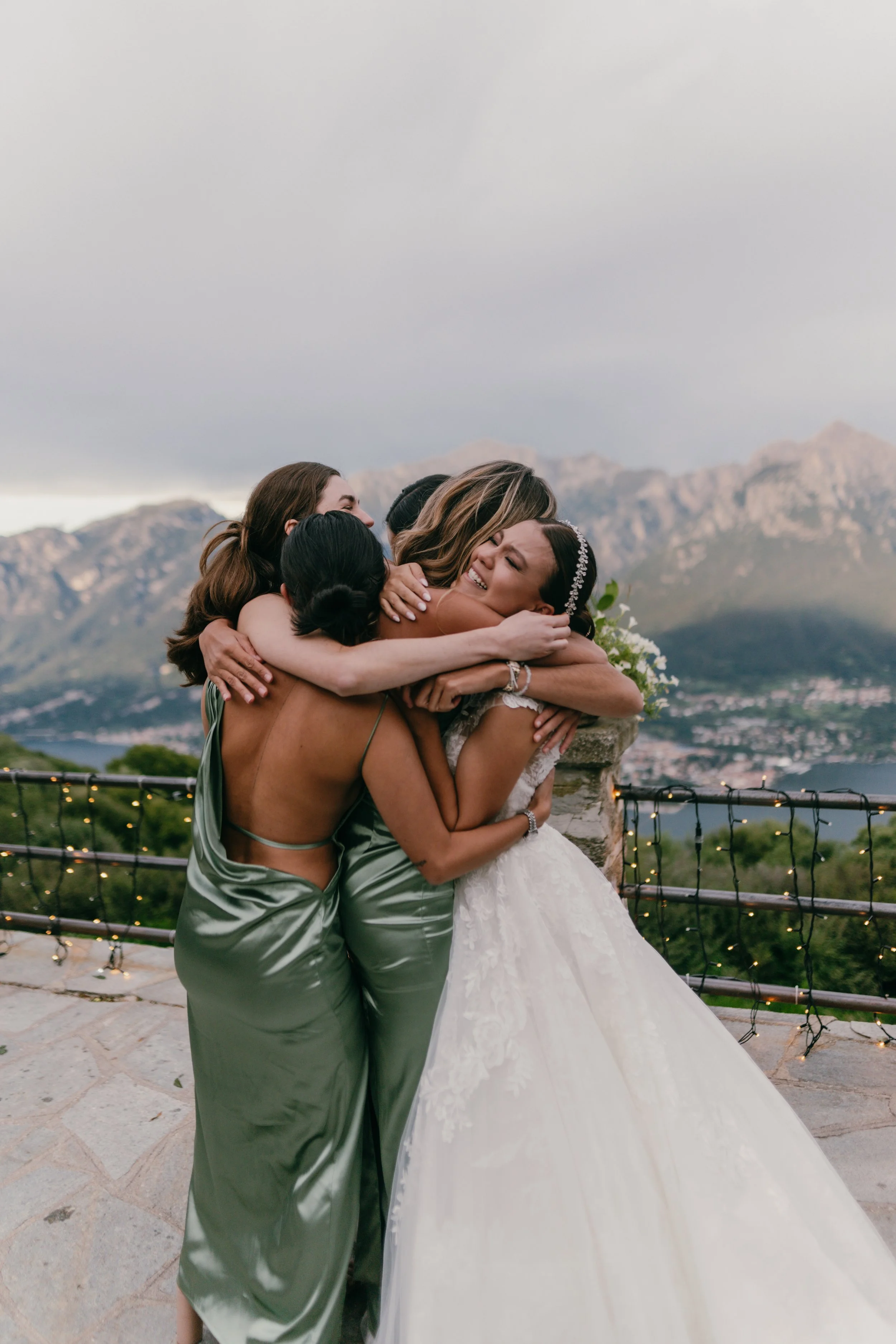 Group of women hugging at a wedding celebration outdoors with mountains and cloudy sky in the background.