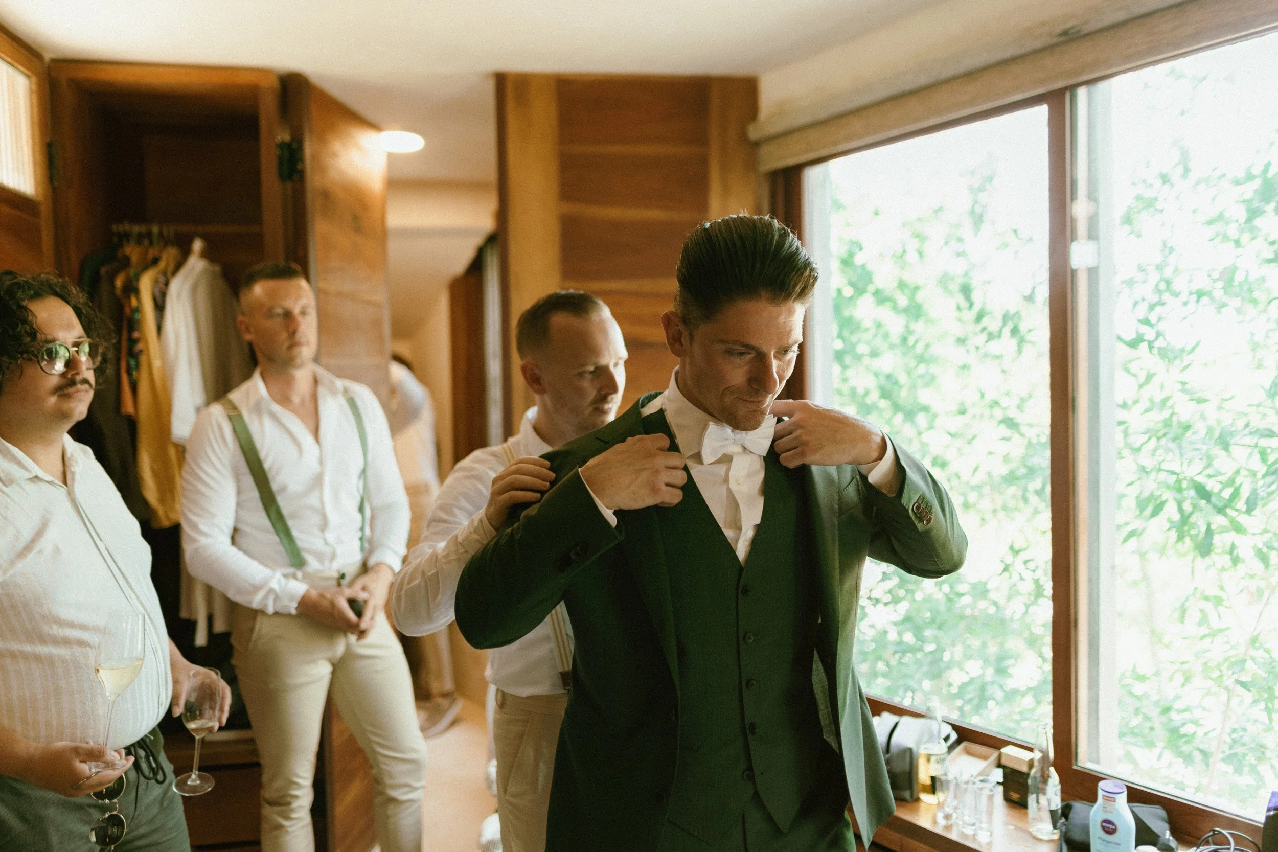 A groom in a green suit adjusts his bow tie as his groomsmen help him prepare in a wood-paneled room with large windows showing greenery outside. The men are wearing white shirts and suspenders, and one is holding a glass of white wine.
