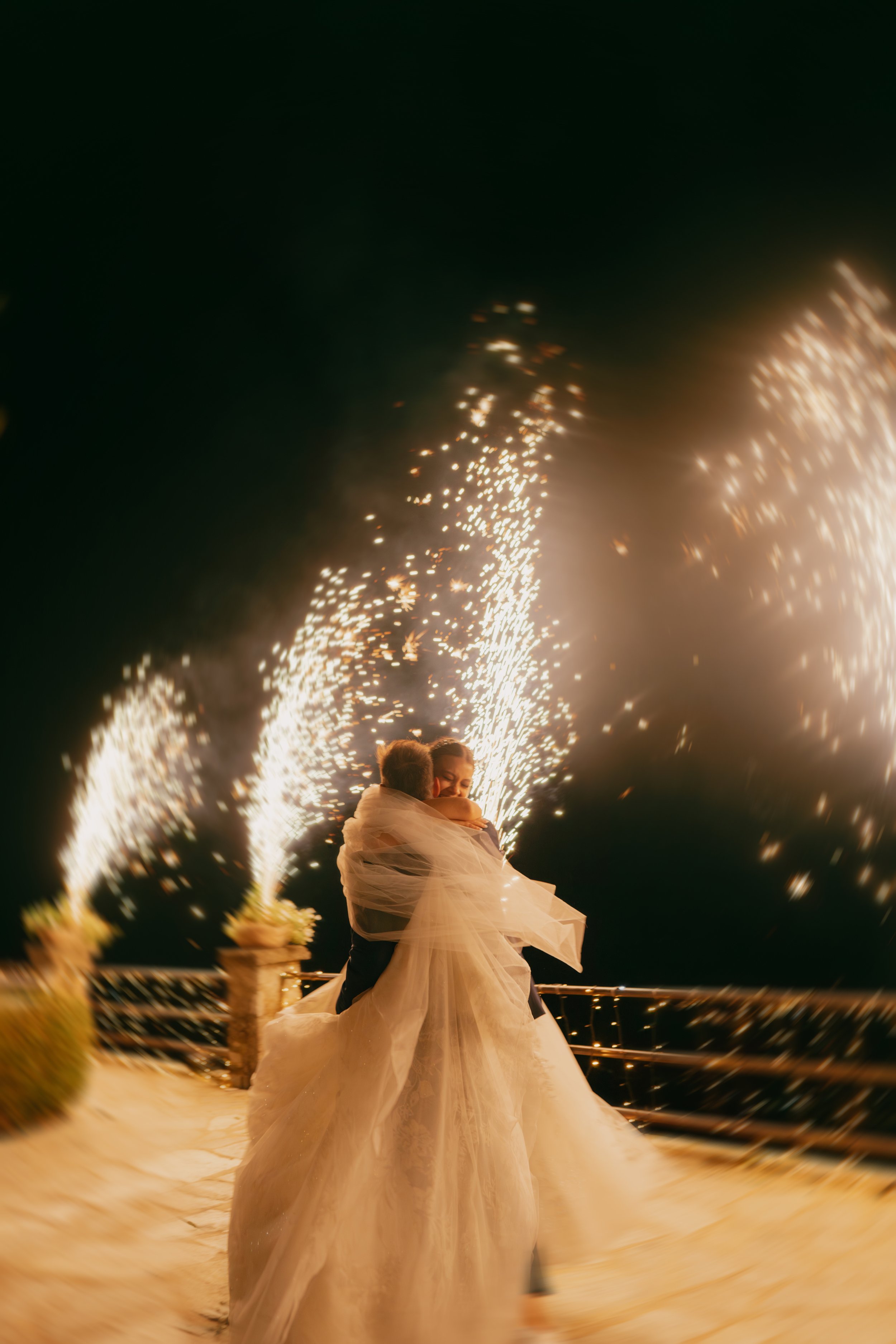 A bride and groom embrace on a dance floor at night with fireworks in the background.