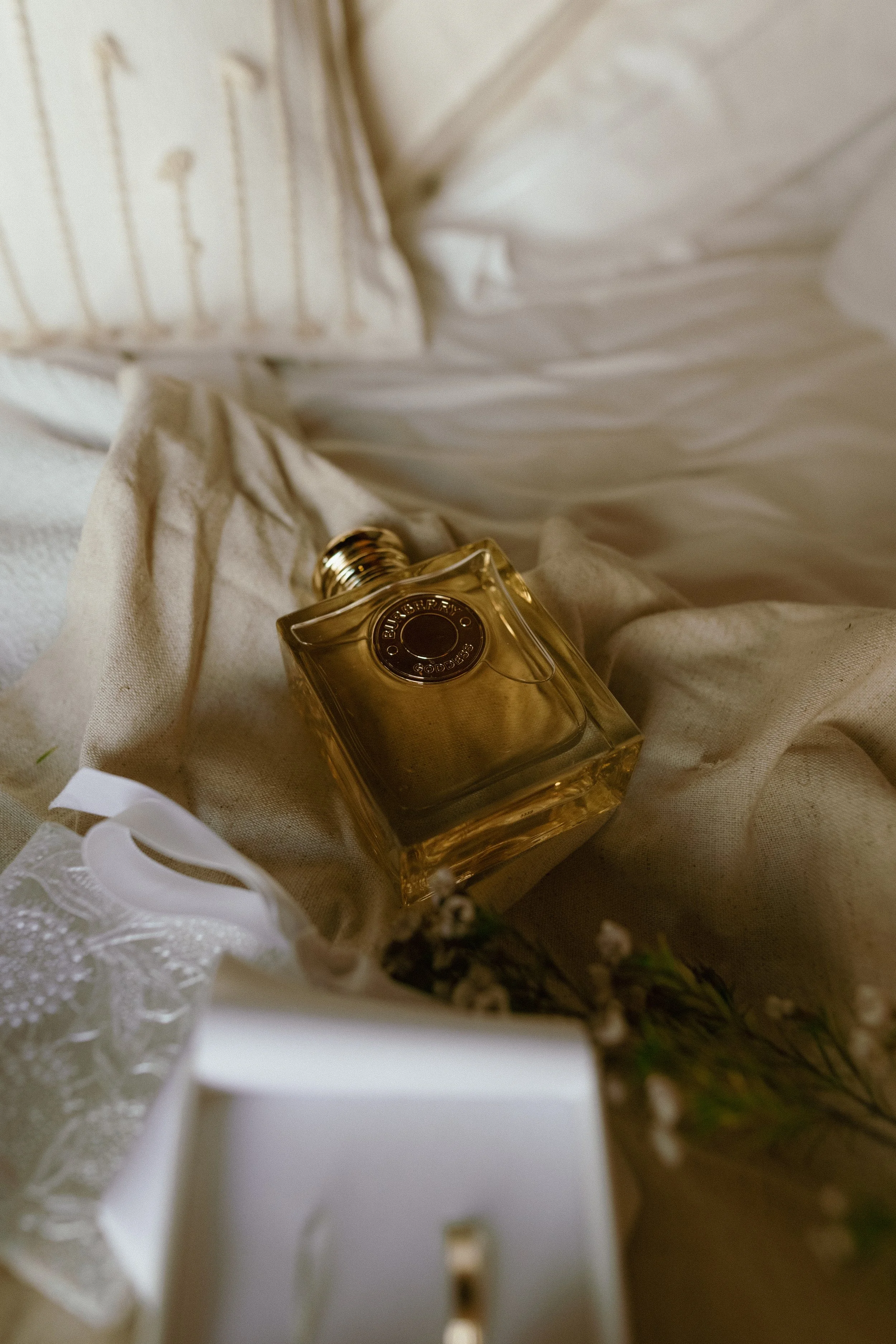 A perfume bottle with a gold cap lying on a beige fabric bed sheet, with a linen pillow and a dried flower nearby.