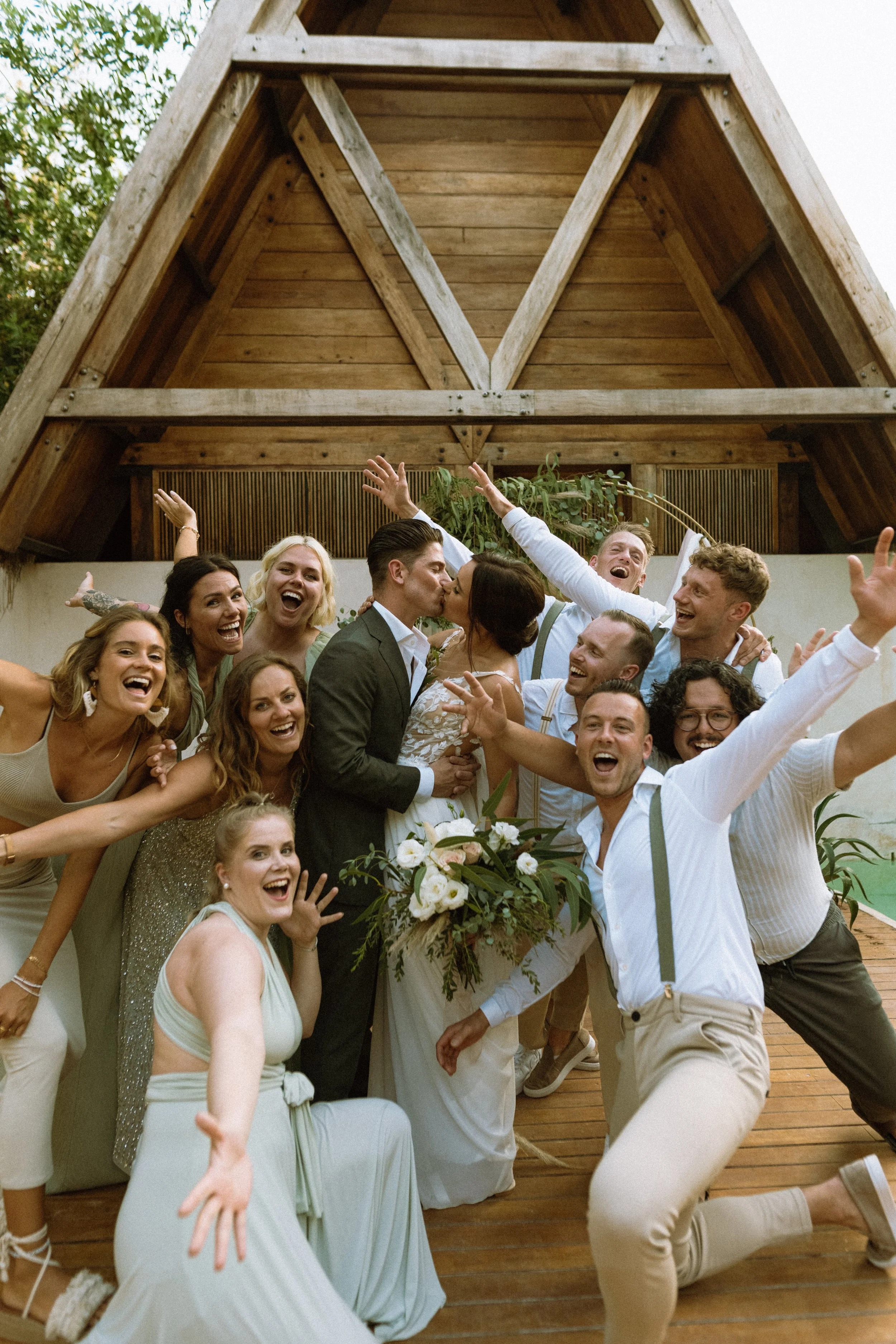 A group of happy people celebrating a wedding, with a bride and groom kissing in the center, all smiling and raising their hands, outside under a wooden roofed structure.