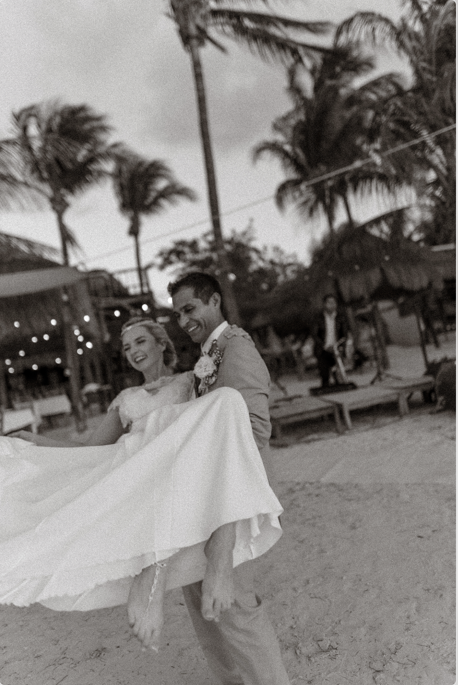 A black-and-white photo of a man in a suit carrying a smiling woman in a wedding dress on a beach with palm trees and huts in the background.