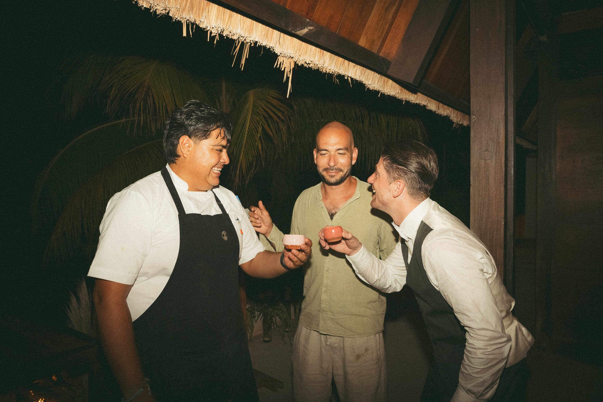 Three men enjoying a moment together at a restaurant or bar, holding small cups and smiling; one man is wearing a chef's uniform, another is in a gray vest and white shirt, and the third man is in a green shirt. Wooden ceiling and tropical plants are