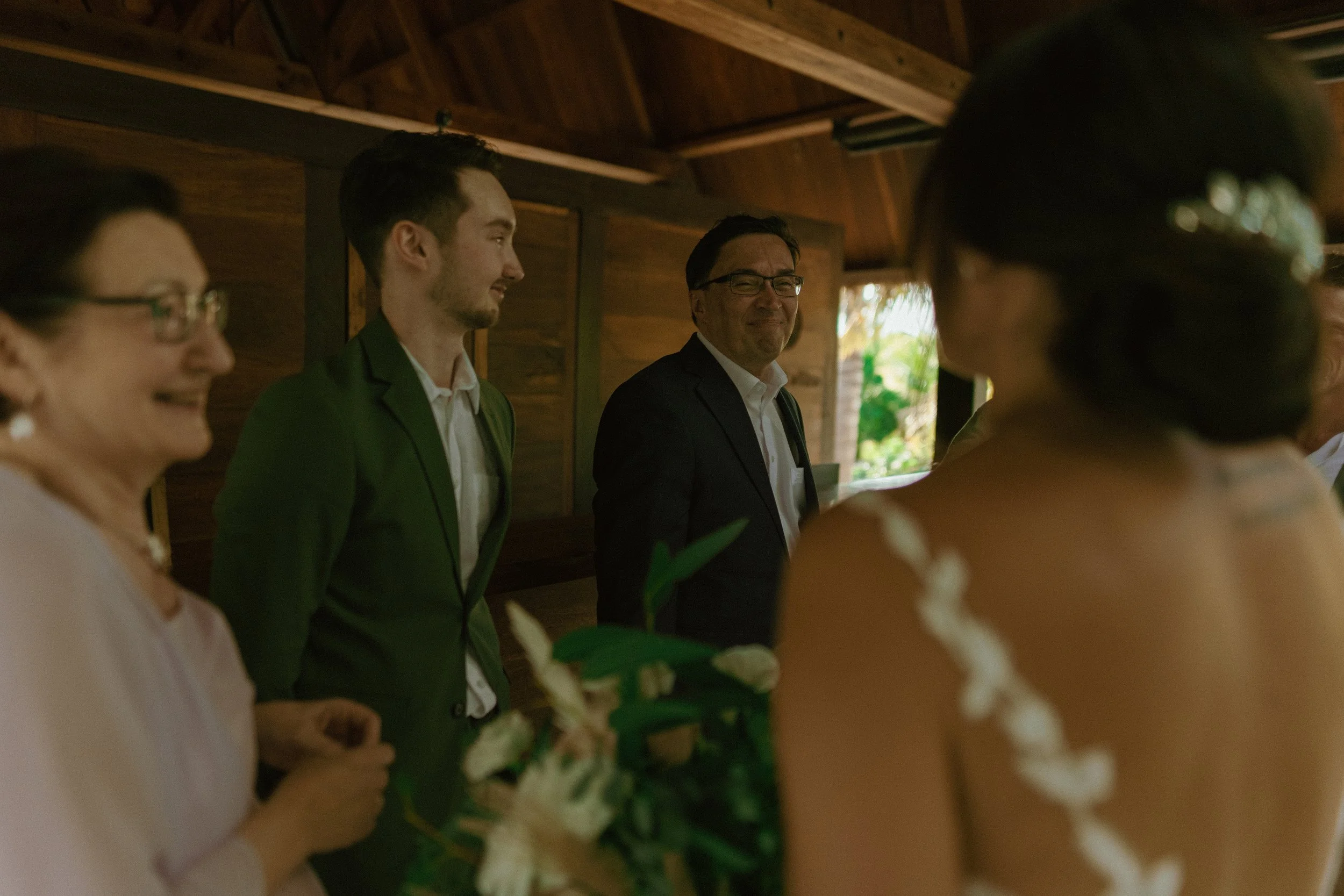 People smiling and talking at a gathering inside a wooden structure, with a woman holding a bouquet of flowers.