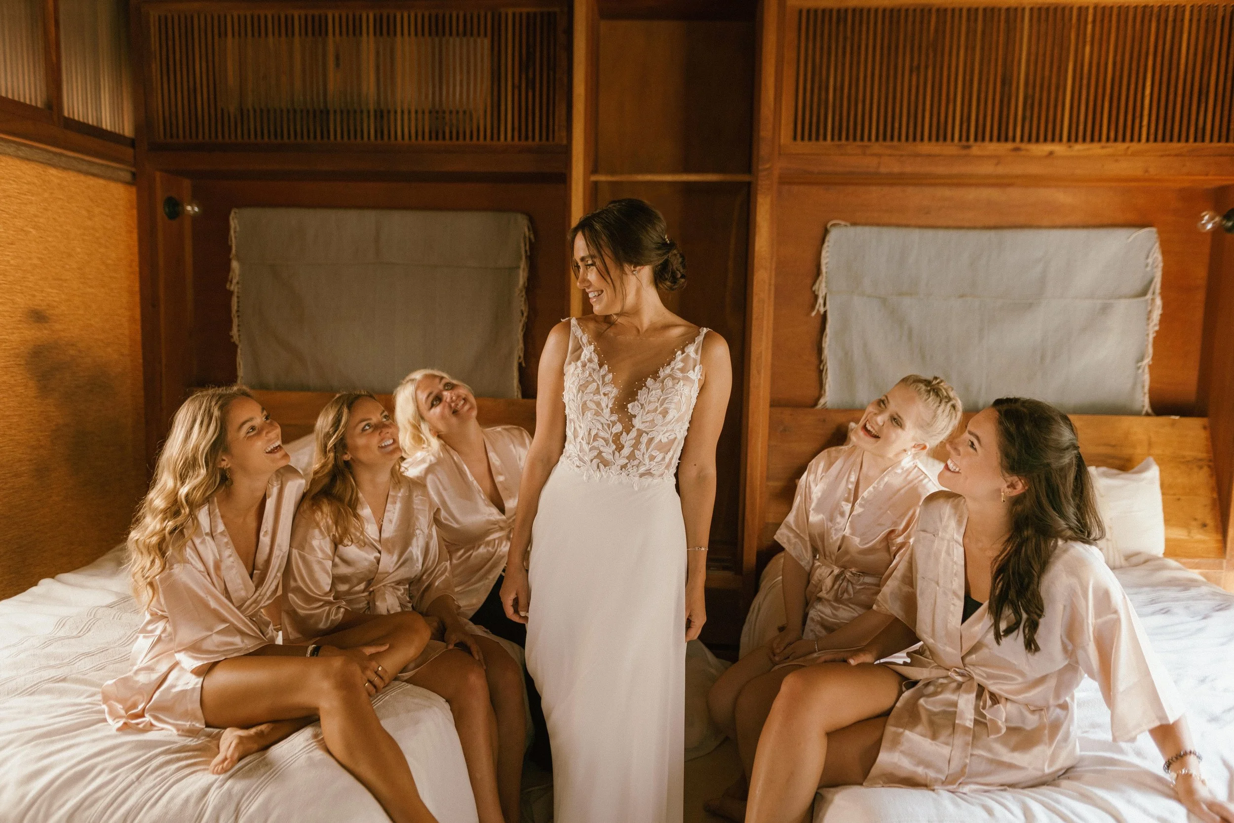 Bride in a white gown standing between five bridesmaids in satin robes, sitting on beds in a wooden room, smiling and looking at her.