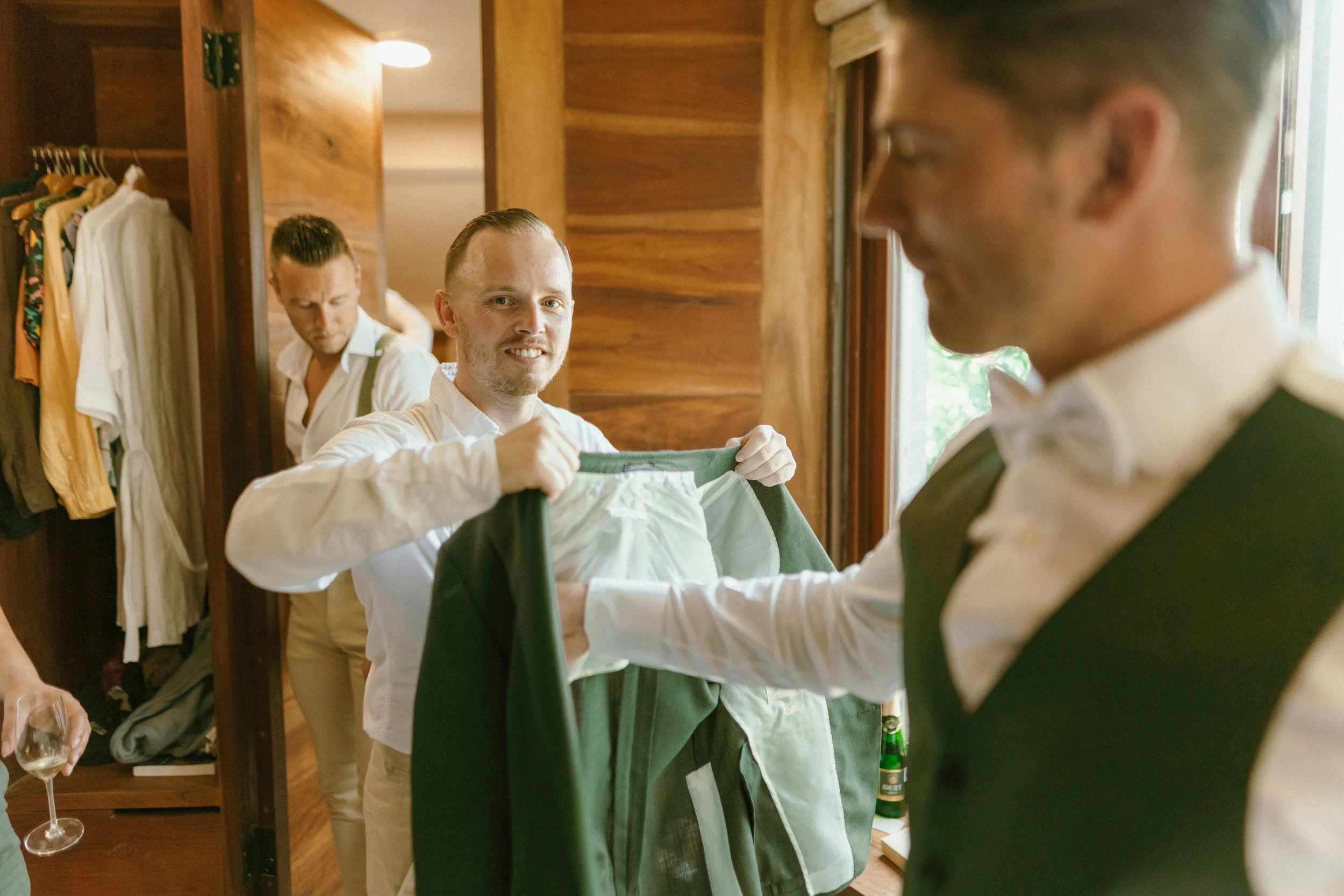 Group of men preparing for a formal event in a wooden-paneled room, with one holding a green jacket and others adjusting clothing, and a hand holding a wine glass visible.