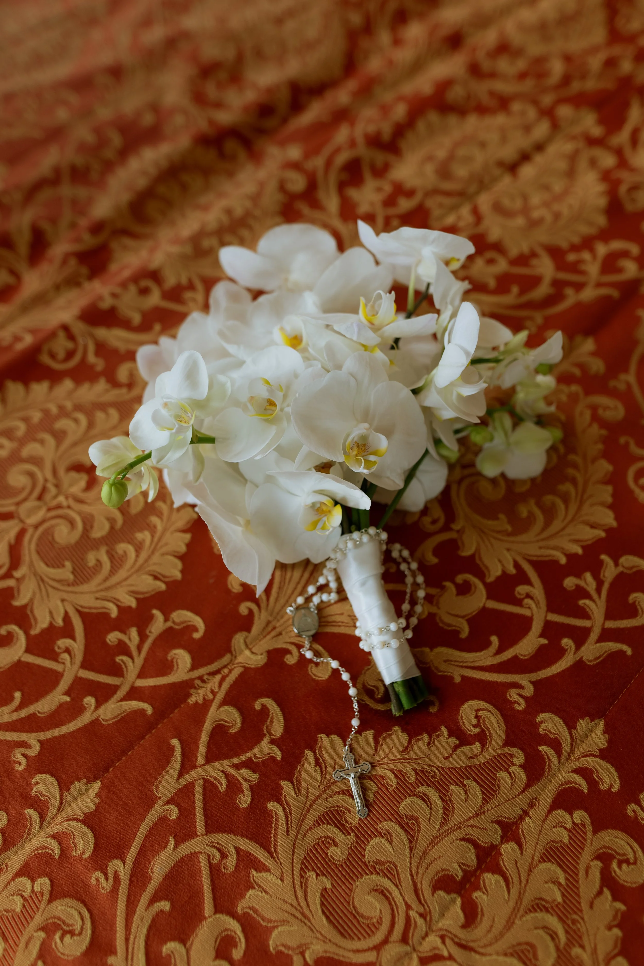 White orchid bouquet with a rosary and a watch on a patterned orange and gold fabric