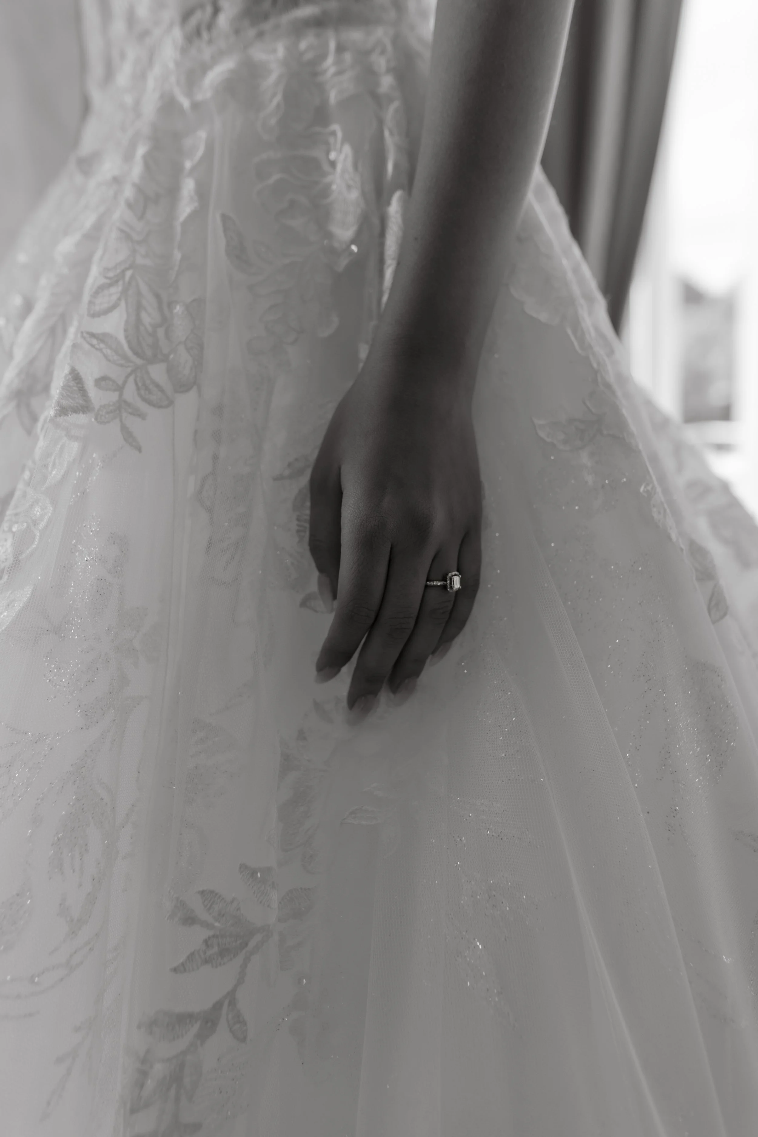 Close-up of a person's hand with an engagement ring on their finger, resting on a wedding dress with floral embroidery.