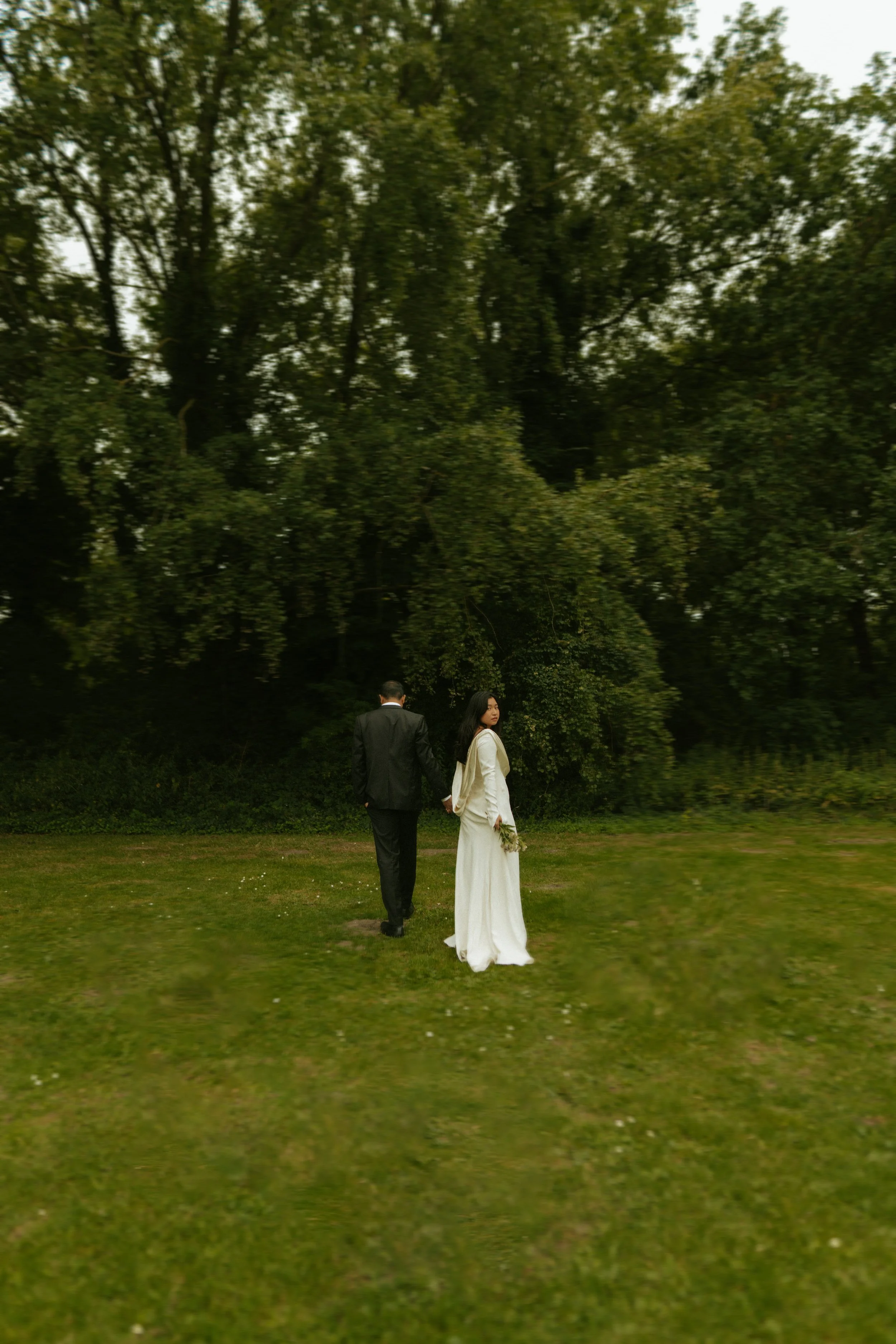 A bride and groom holding hands walking away in a grassy area with large trees in the background.
