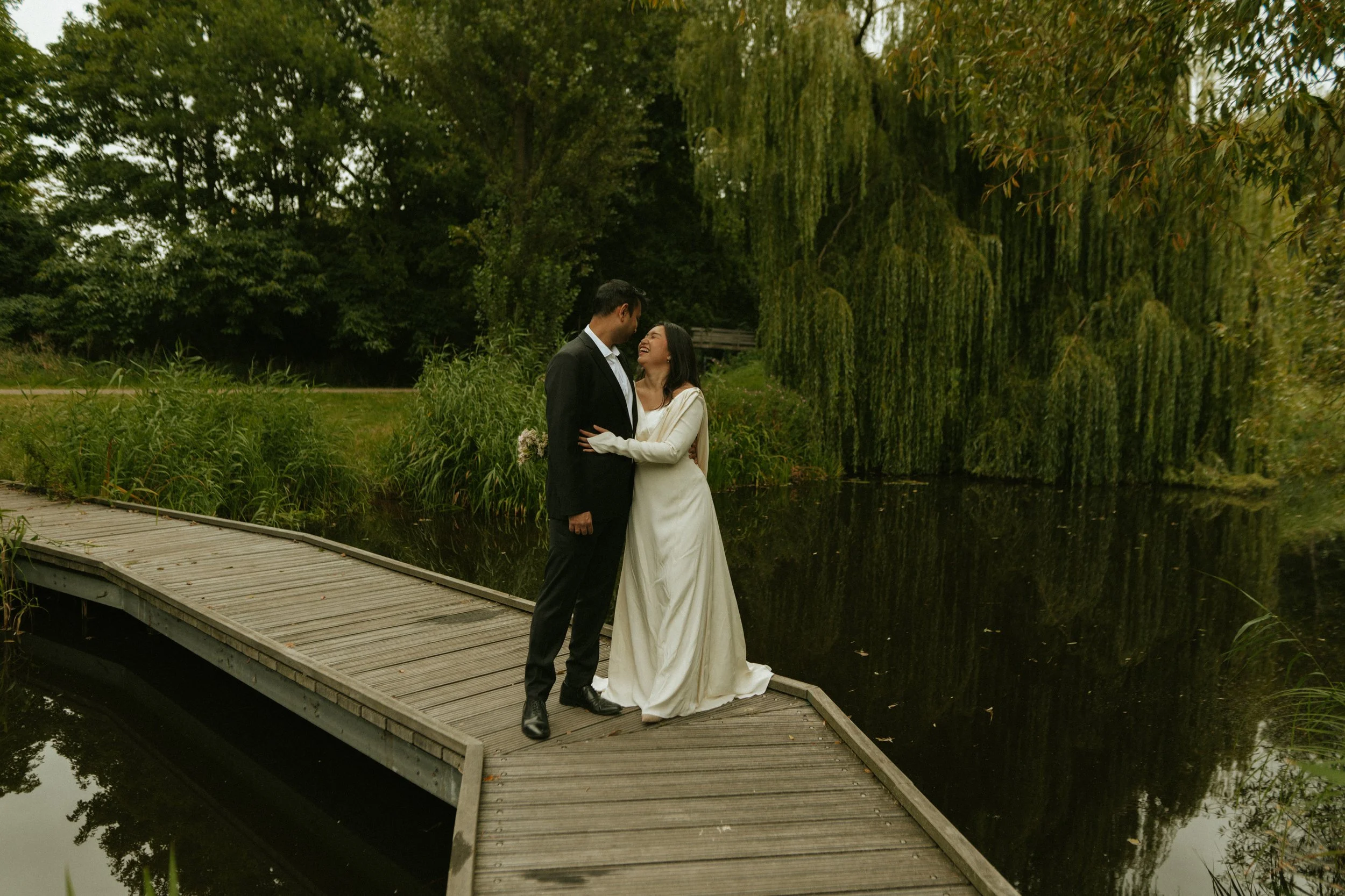 A newlywed couple stands on a wooden pathway by a pond in a lush, green park. The bride is wearing a long white dress, and the groom is in a black suit. They are smiling and gazing at each other.