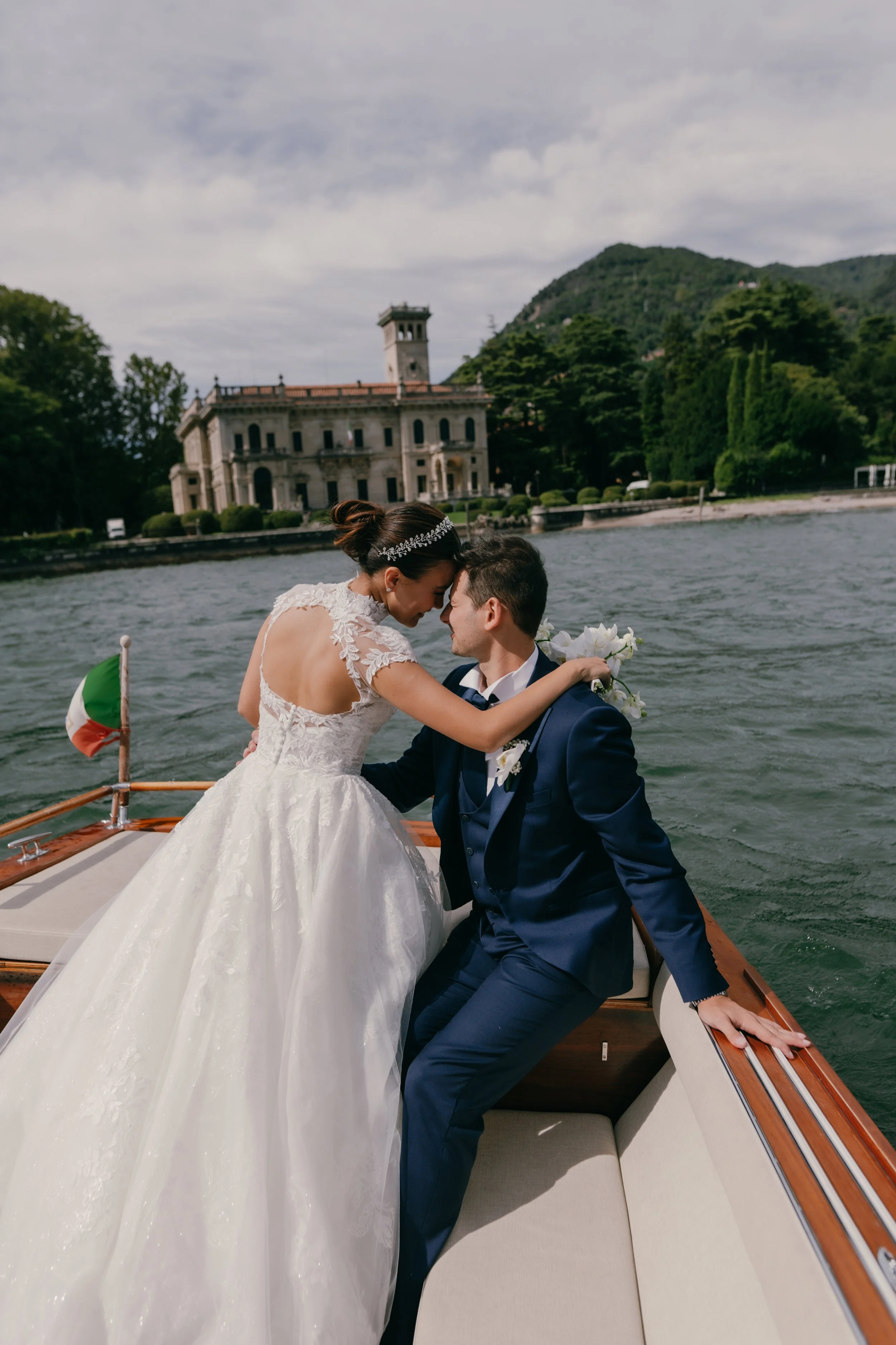 Bride and groom sharing a close, intimate moment on a boat with a historic building and lush green hills in the background.