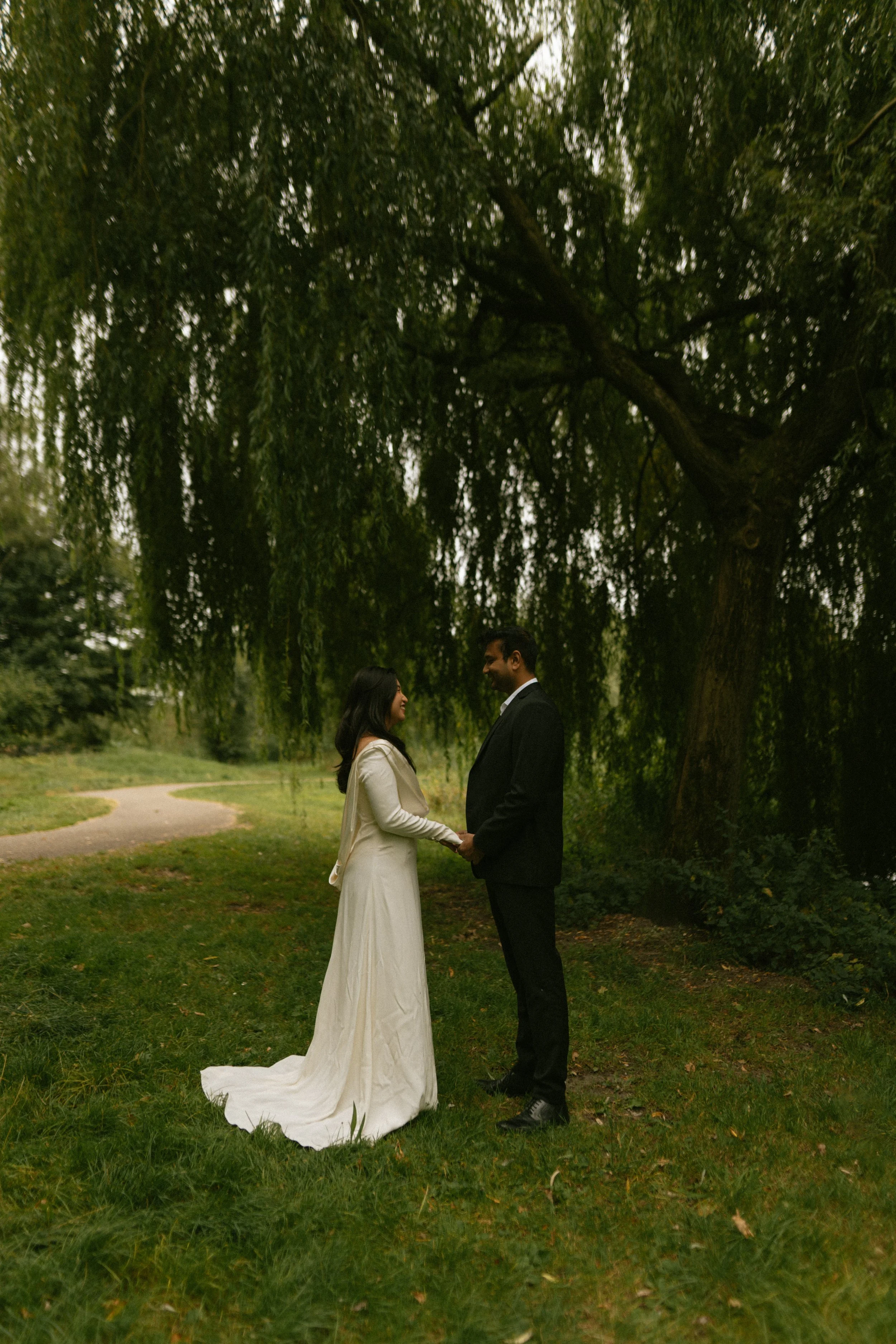 A couple holding hands and looking at each other under a large green tree in a park.
