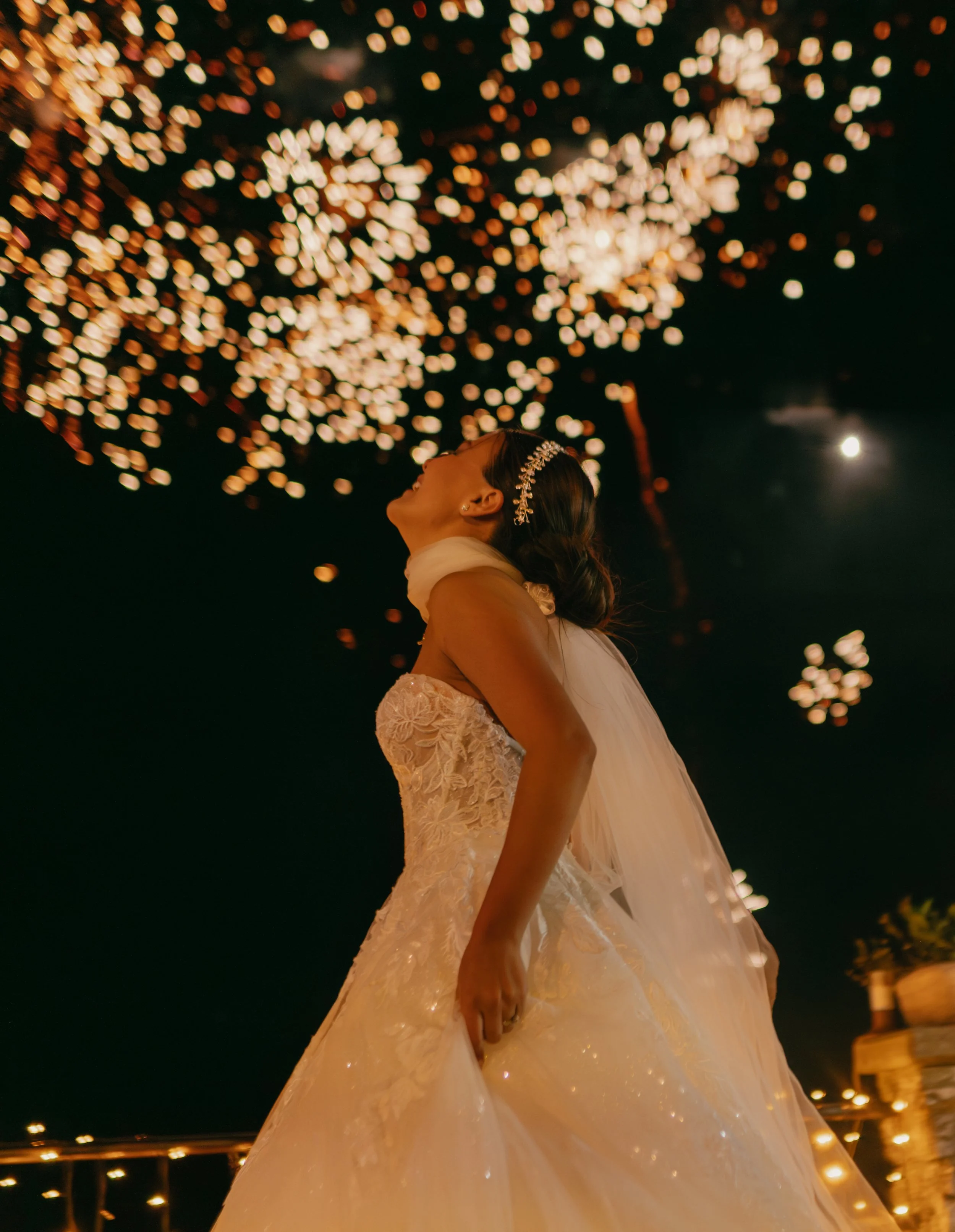 A bride in a lace wedding gown and veil watching fireworks at night.