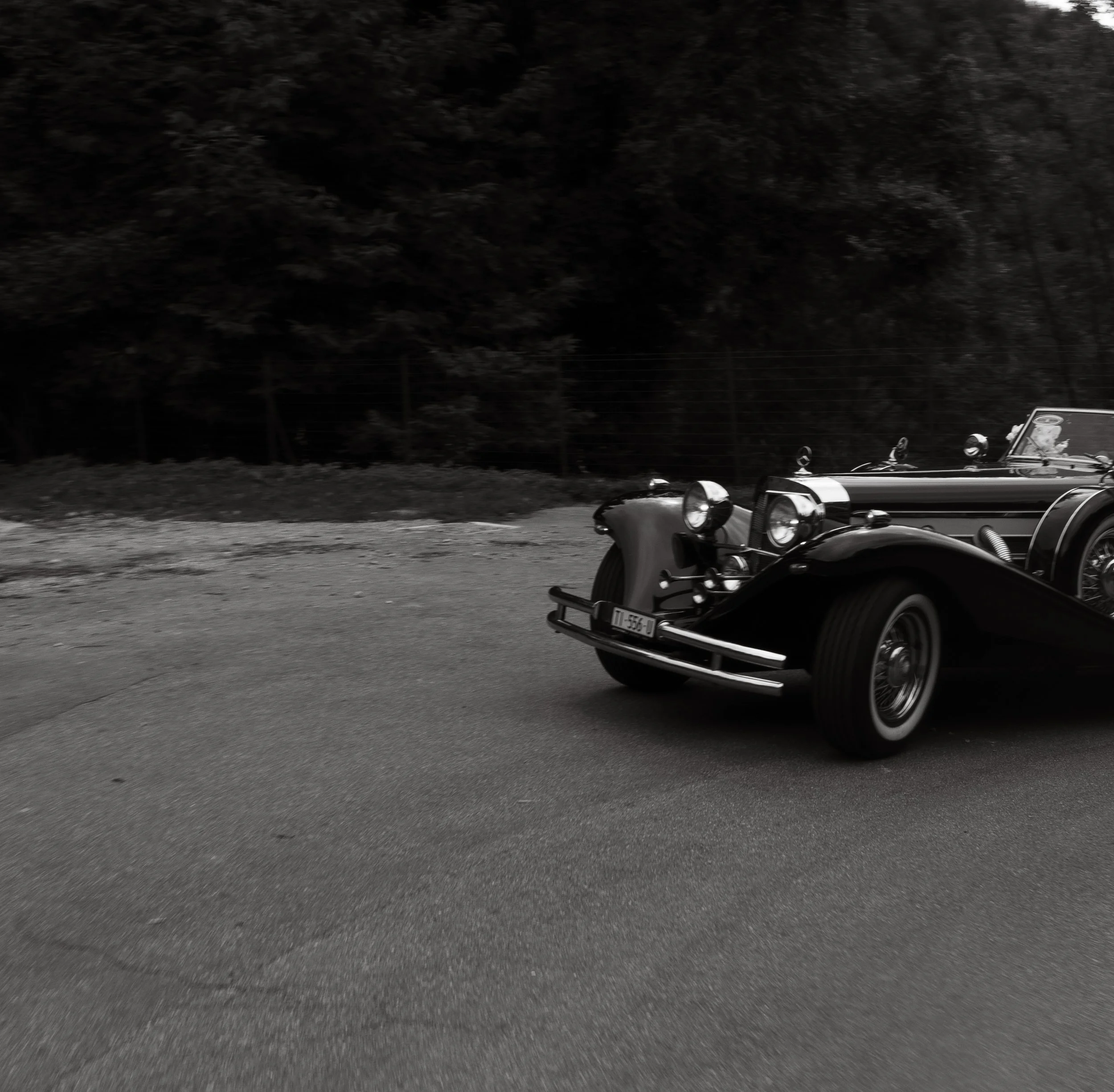 A vintage black convertible car with round headlights driving on a paved road with a wooded background.