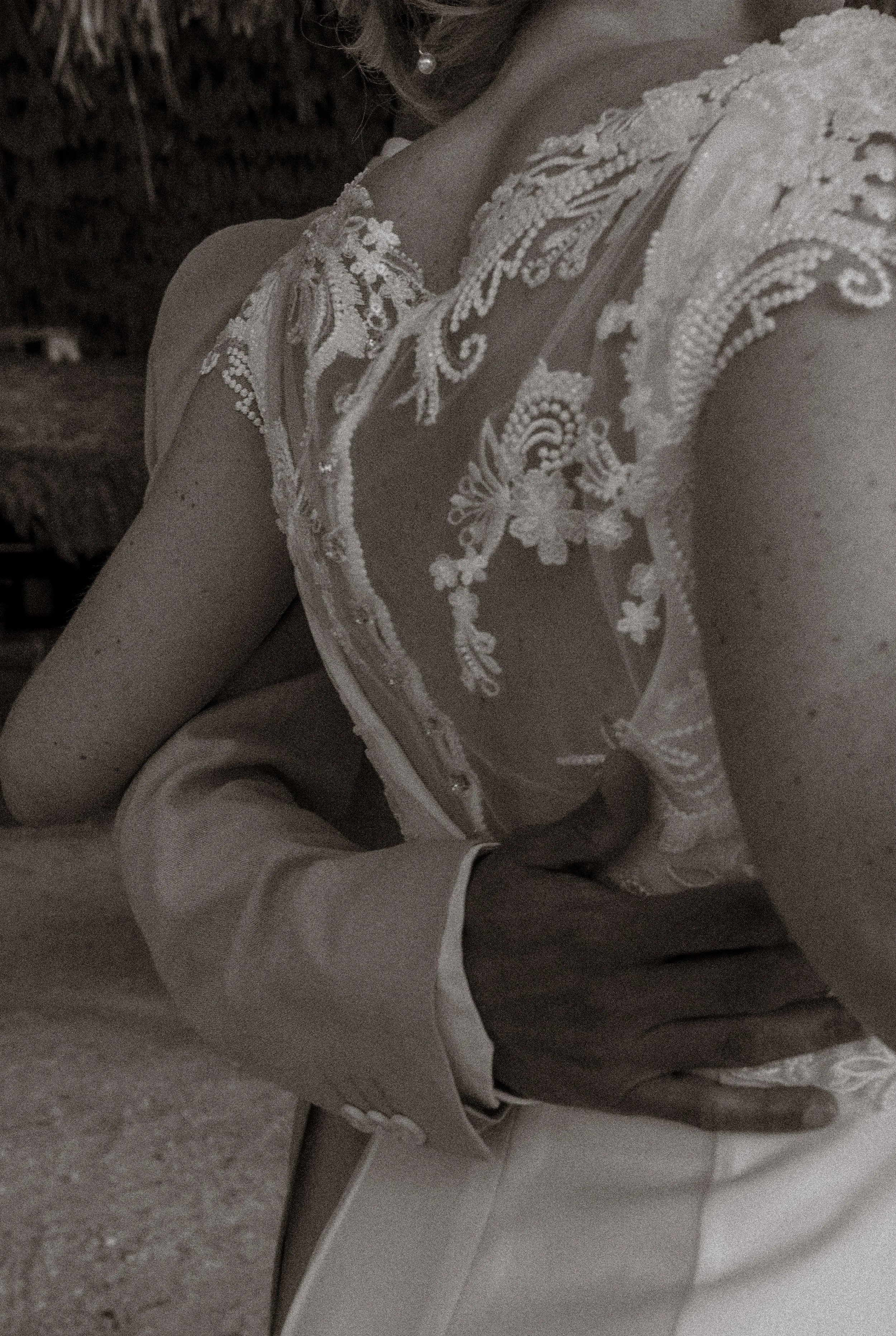 A person wearing an elegant wedding dress with intricate lace, and a person in a tuxedo holding them from behind, outdoors during the daytime.