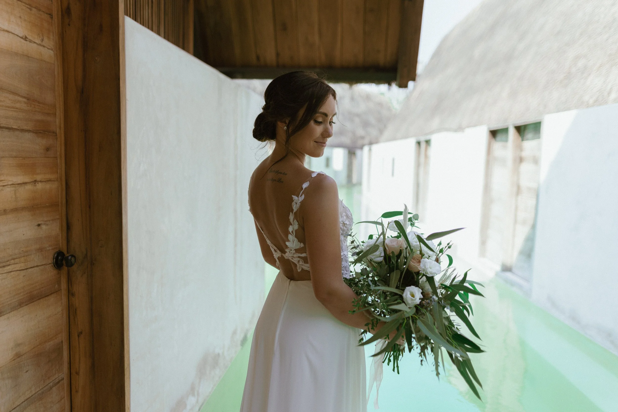 A bride in a wedding dress holding a bouquet of white and blush flowers with greenery, standing in a modern, minimalist hallway with wooden and white walls and a green floor.