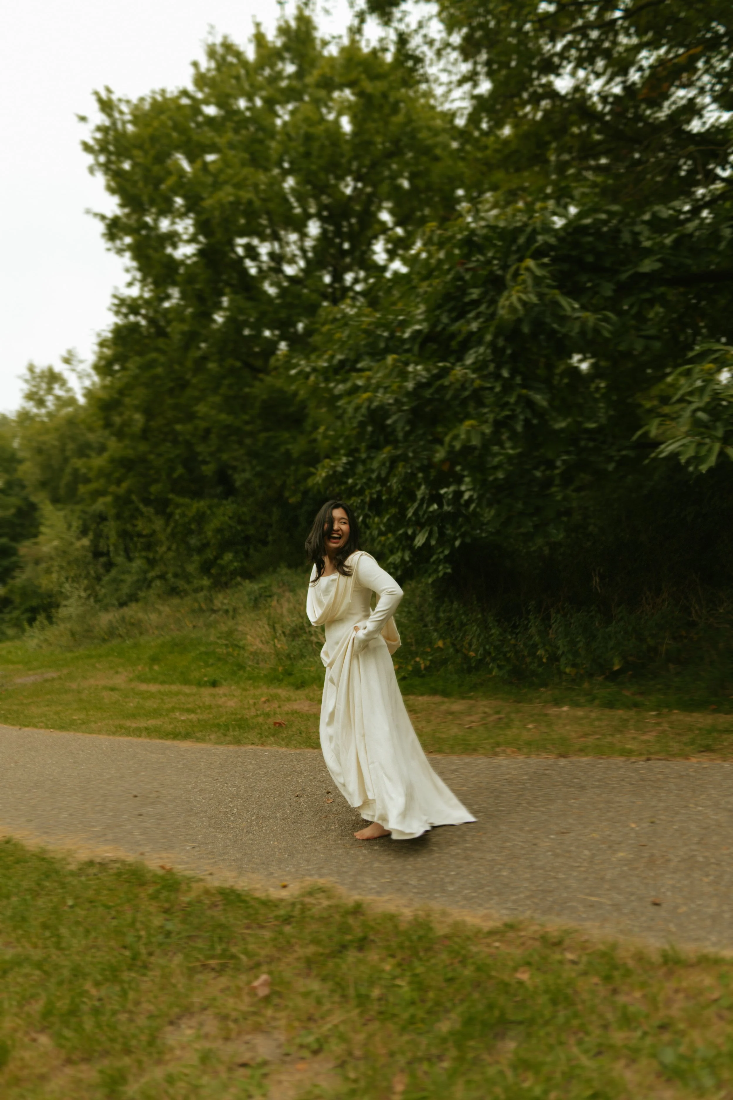 A woman in a white dress standing on a path outdoors, smiling and laughing, with large green trees in the background.