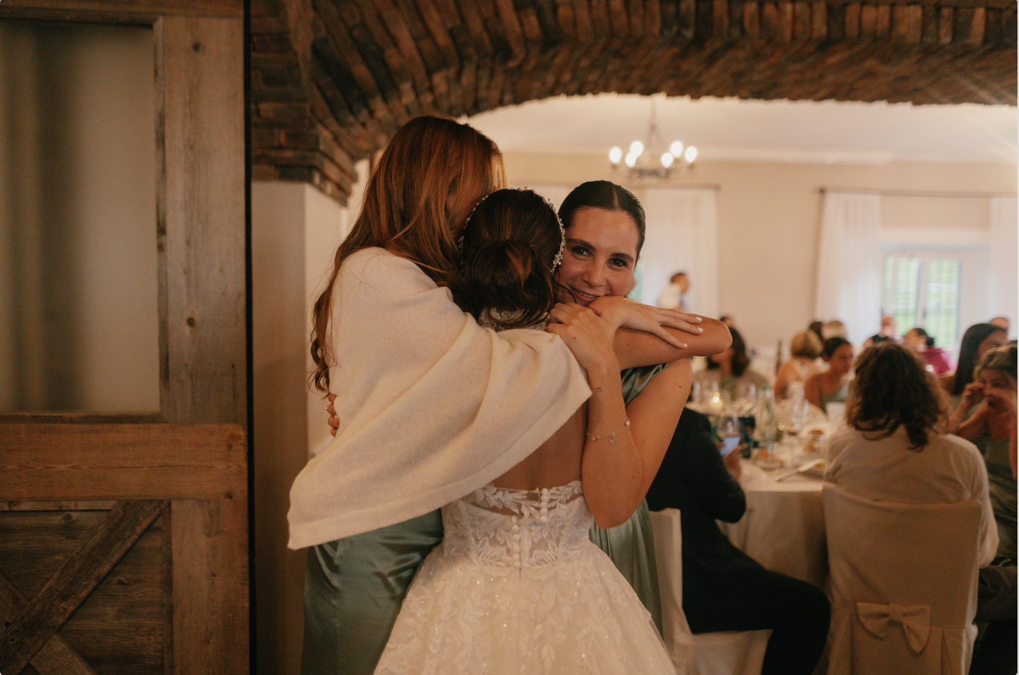 A bride in a wedding dress hugging two women, one with red hair and the other with brown hair, at a wedding reception with guests seated at tables in the background.