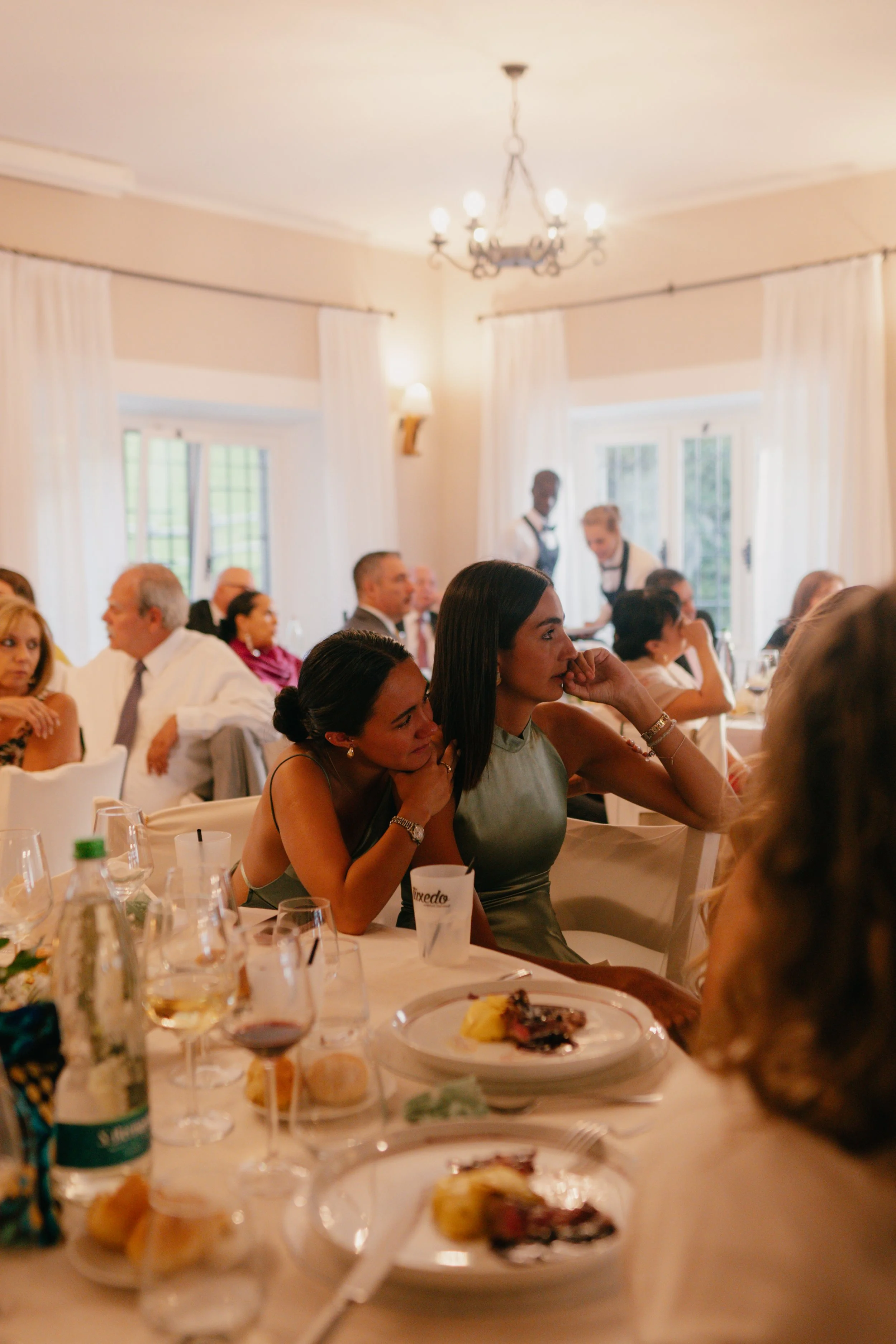 People sitting at a banquet table listening to a speaker, with food and drinks on the table, in a well-lit dining room with chandeliers and white curtains.