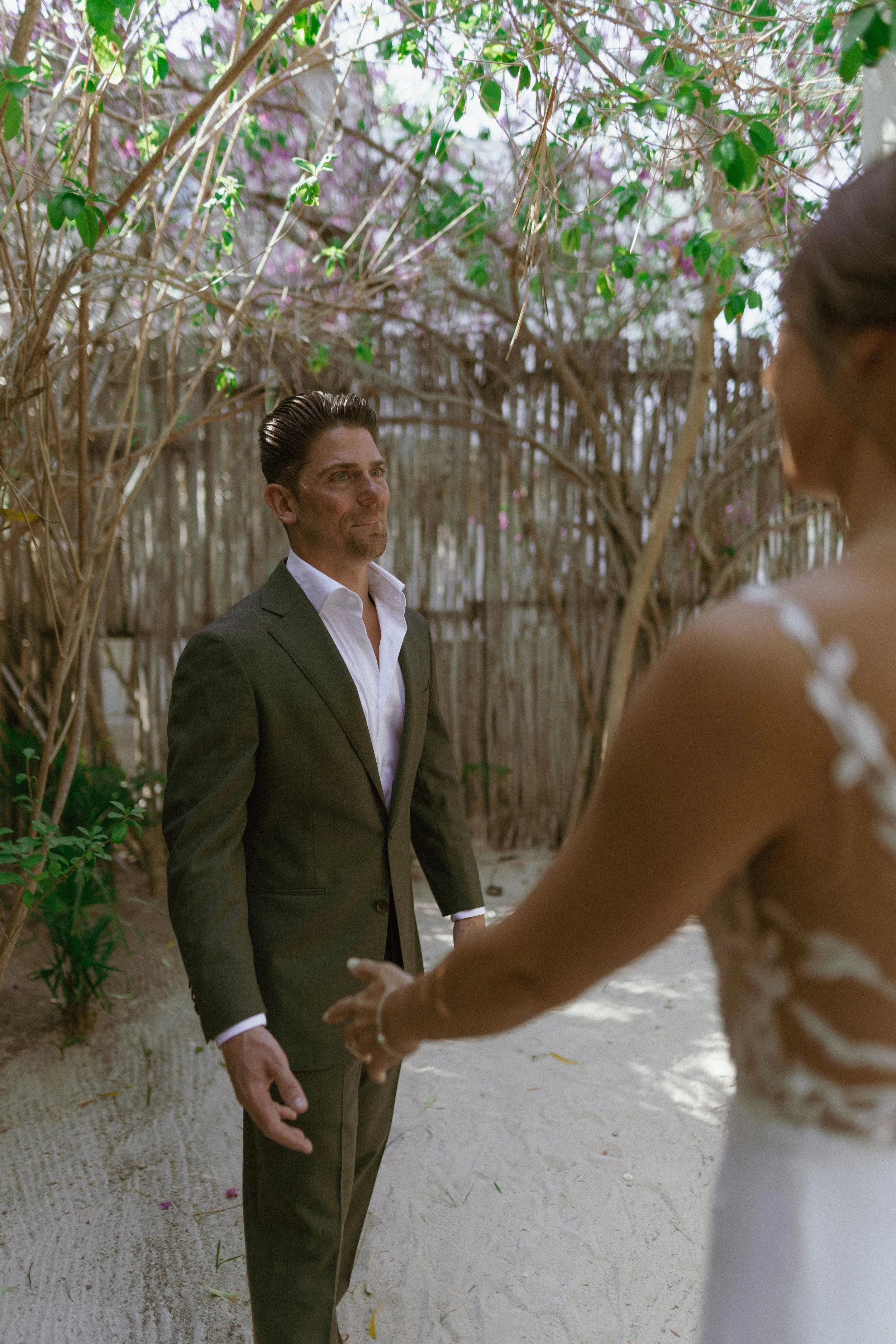 A couple holding hands during their wedding ceremony in a lush outdoor setting with trees and pink flowers.