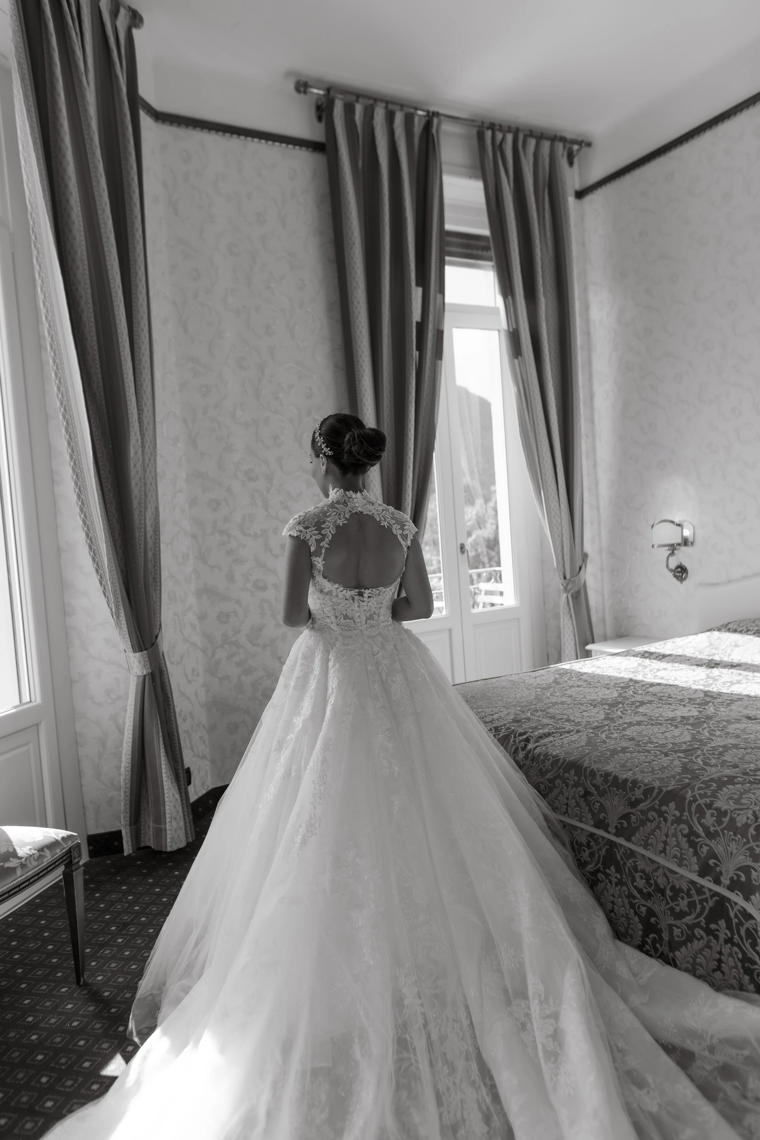 A bride in a wedding dress standing in a elegant hotel room with floral wallpaper, large windows, and heavy drapes.