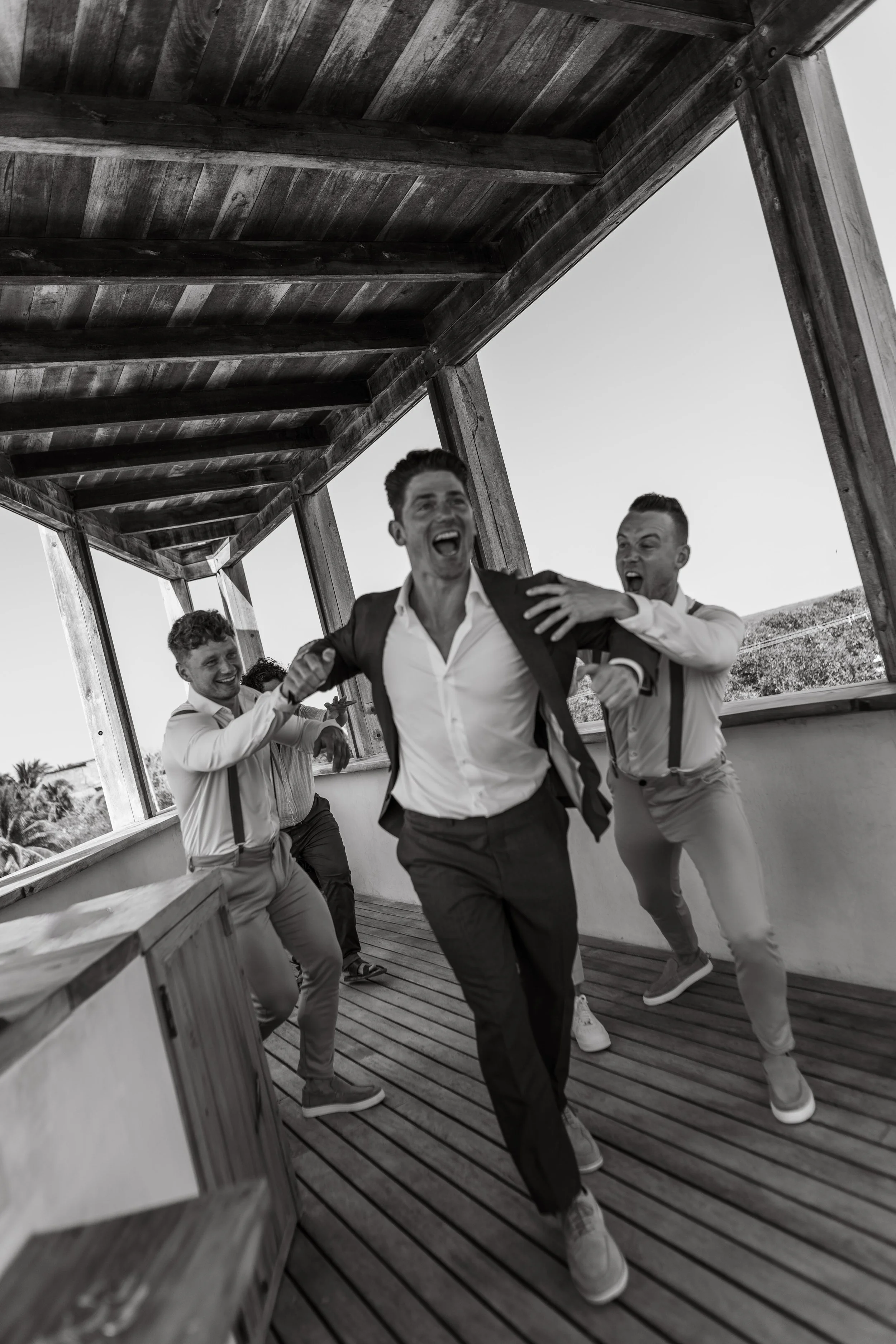A group of men in formal attire having fun and chasing each other on a wooden deck under a covered outdoor area.