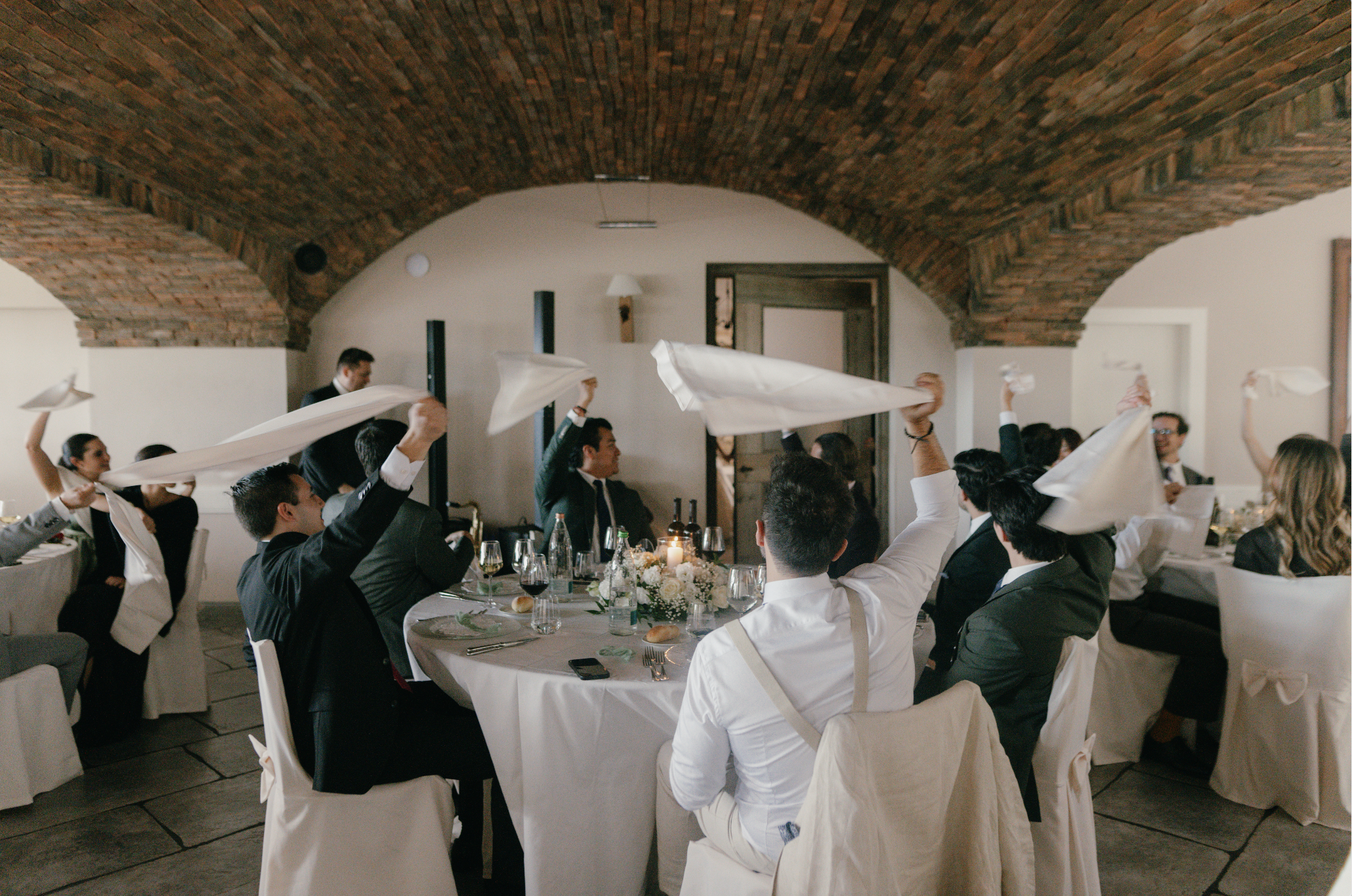 Group of people dressed in formal attire enjoying a celebration or event, sitting around a decorated table with white tablecloth, flowers, and glasses, in a room with brick arched ceiling, some waving white napkins or cloths.