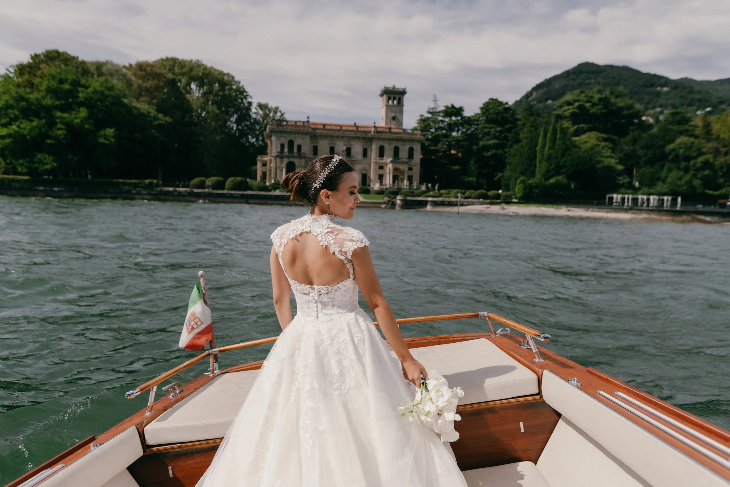 A bride in a white wedding dress holding a bouquet of white flowers, standing on a boat on a lake with a historic building and green trees in the background.