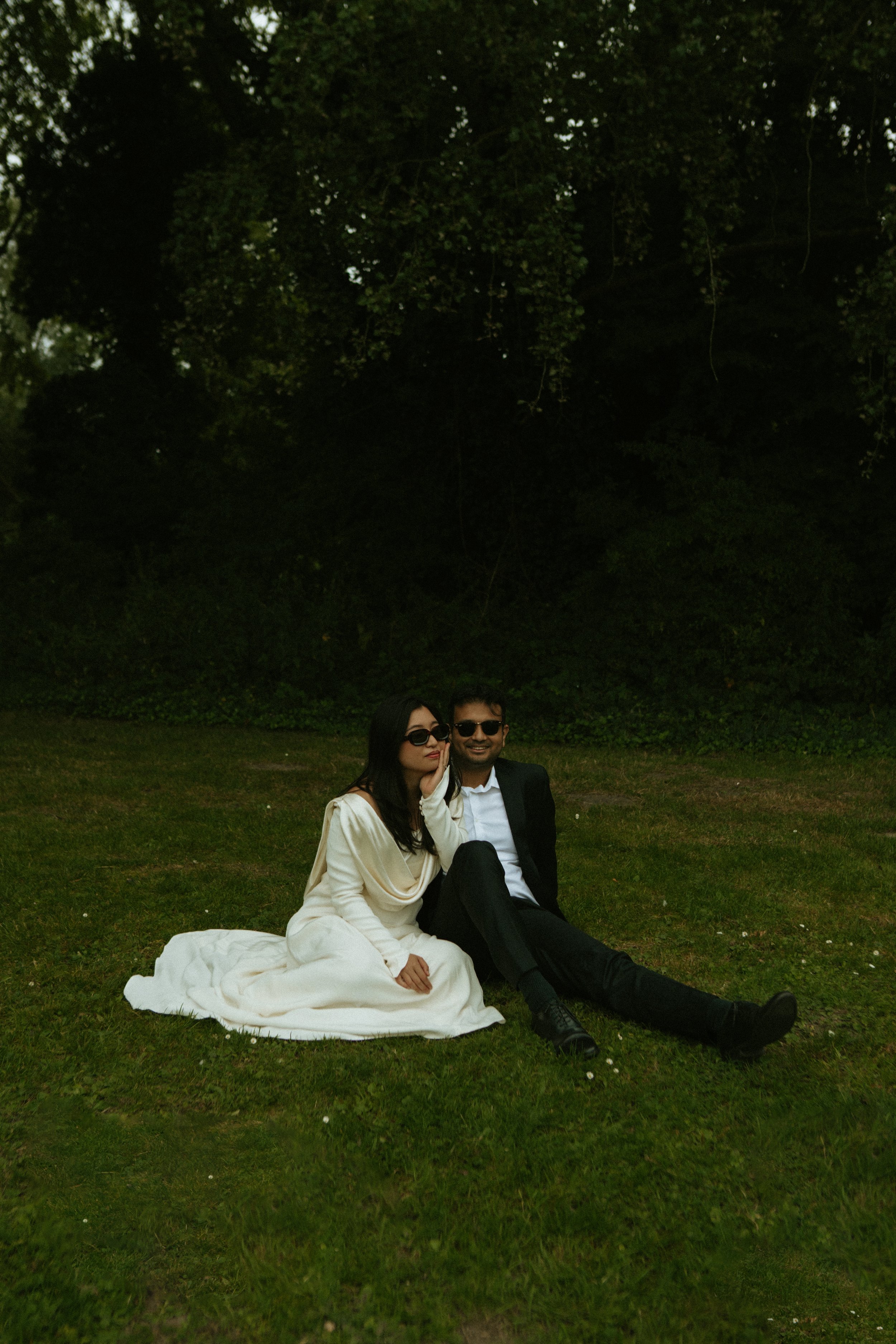 A man and woman sitting on the grass in front of a dark, wooded area. The woman is wearing a white dress and sunglasses, and the man is in a black suit with sunglasses. They appear relaxed and happy.