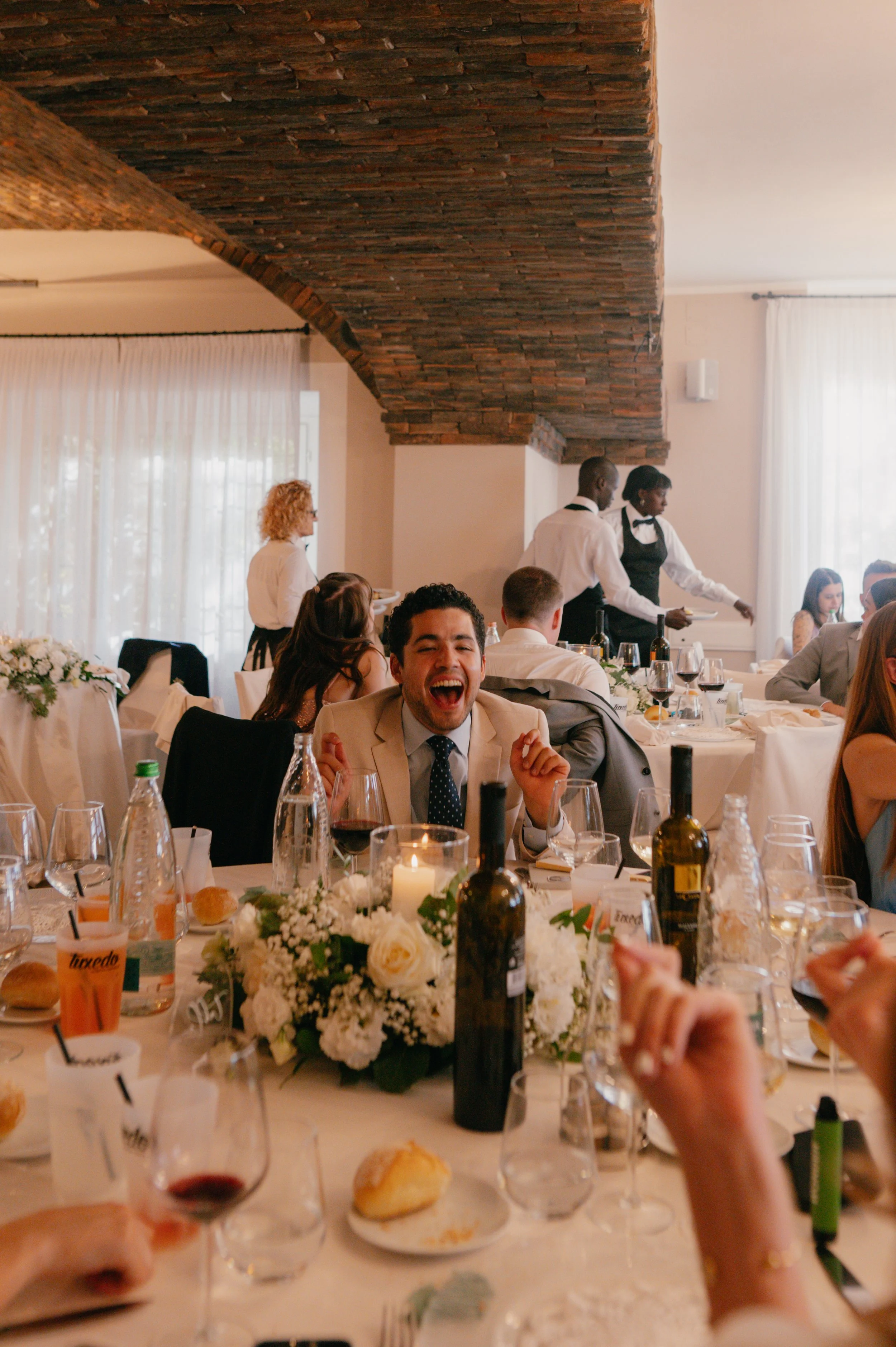 A joyful man laughing at a wedding reception, seated at a table decorated with white flowers, wine bottles, and glasses, surrounded by other guests.