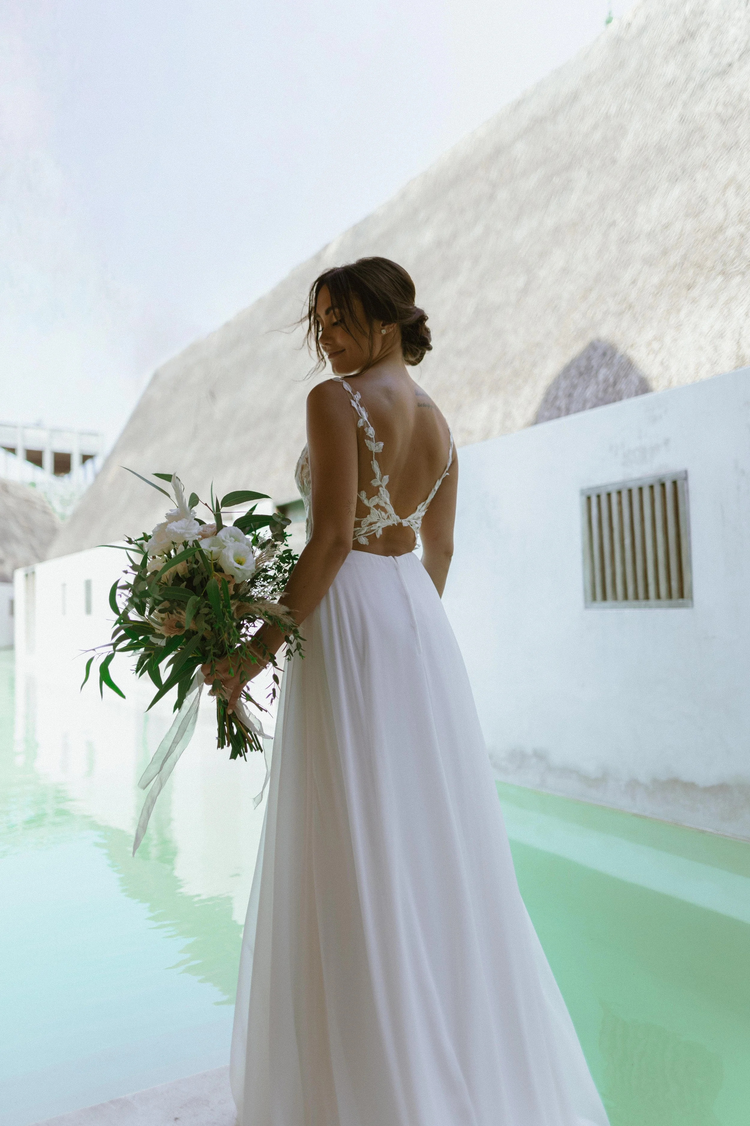 A bride in a white wedding dress holding a bouquet of white and pink flowers, standing near a pool with a white wall and rock formations in the background.