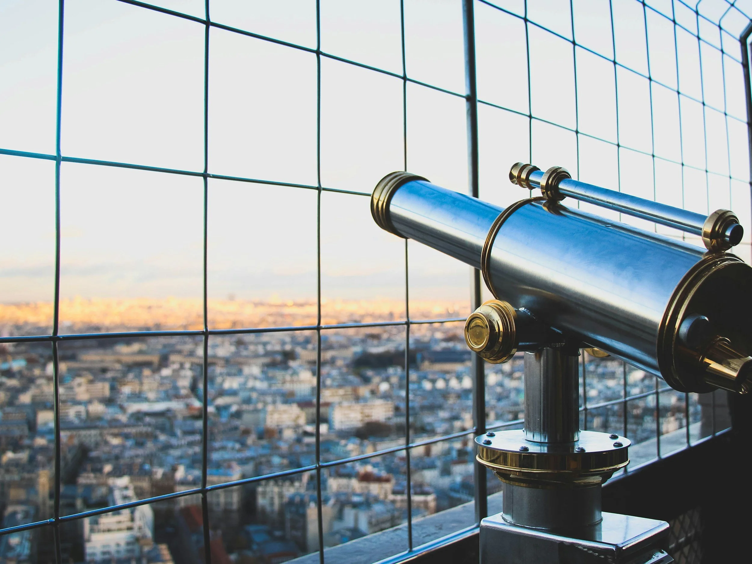 A brass and silver telescope mounted on a stand, looking out over a cityscape through a wire mesh fence during sunset.