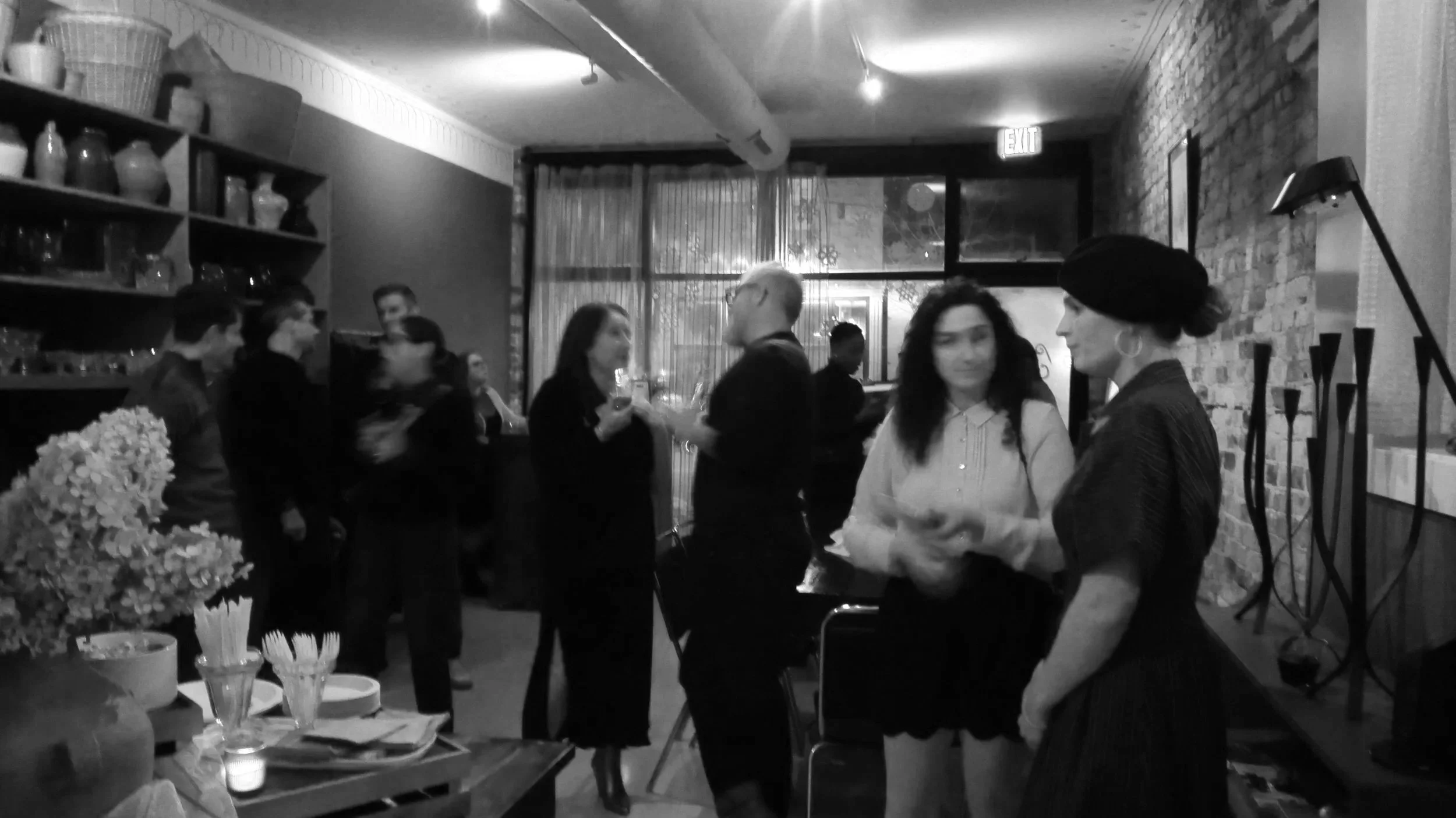 People socializing at a bar or restaurant with shelves of dishes and decorative items, exposed brick wall, and a large window in the background.