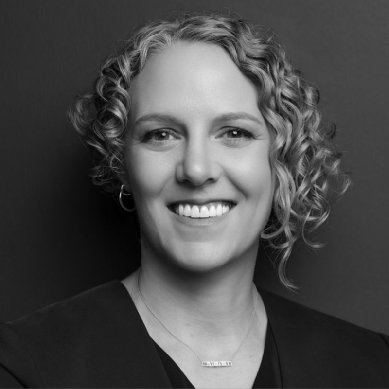 A black and white portrait of a woman with curly blonde hair, smiling, wearing hoop earrings and a necklace, against a dark background.