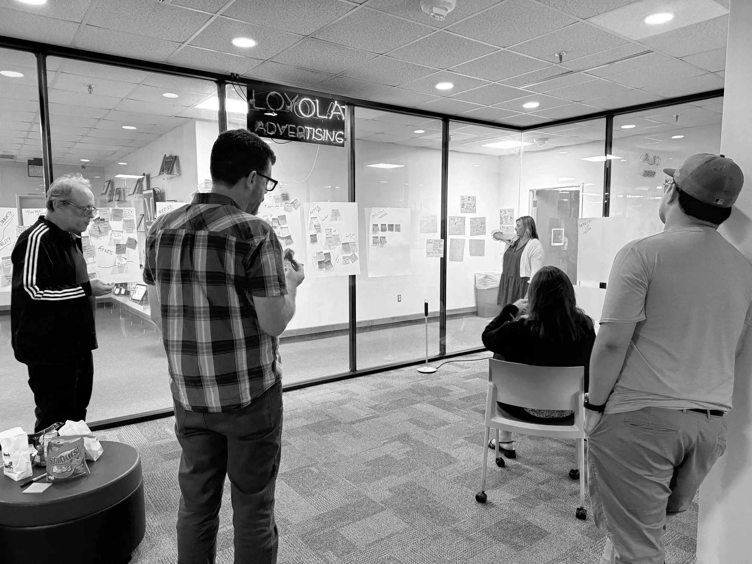 People participating in a brainstorming or planning session behind a glass partition with charts and sketches, while a woman in a dress and white jacket speaks in front of them.